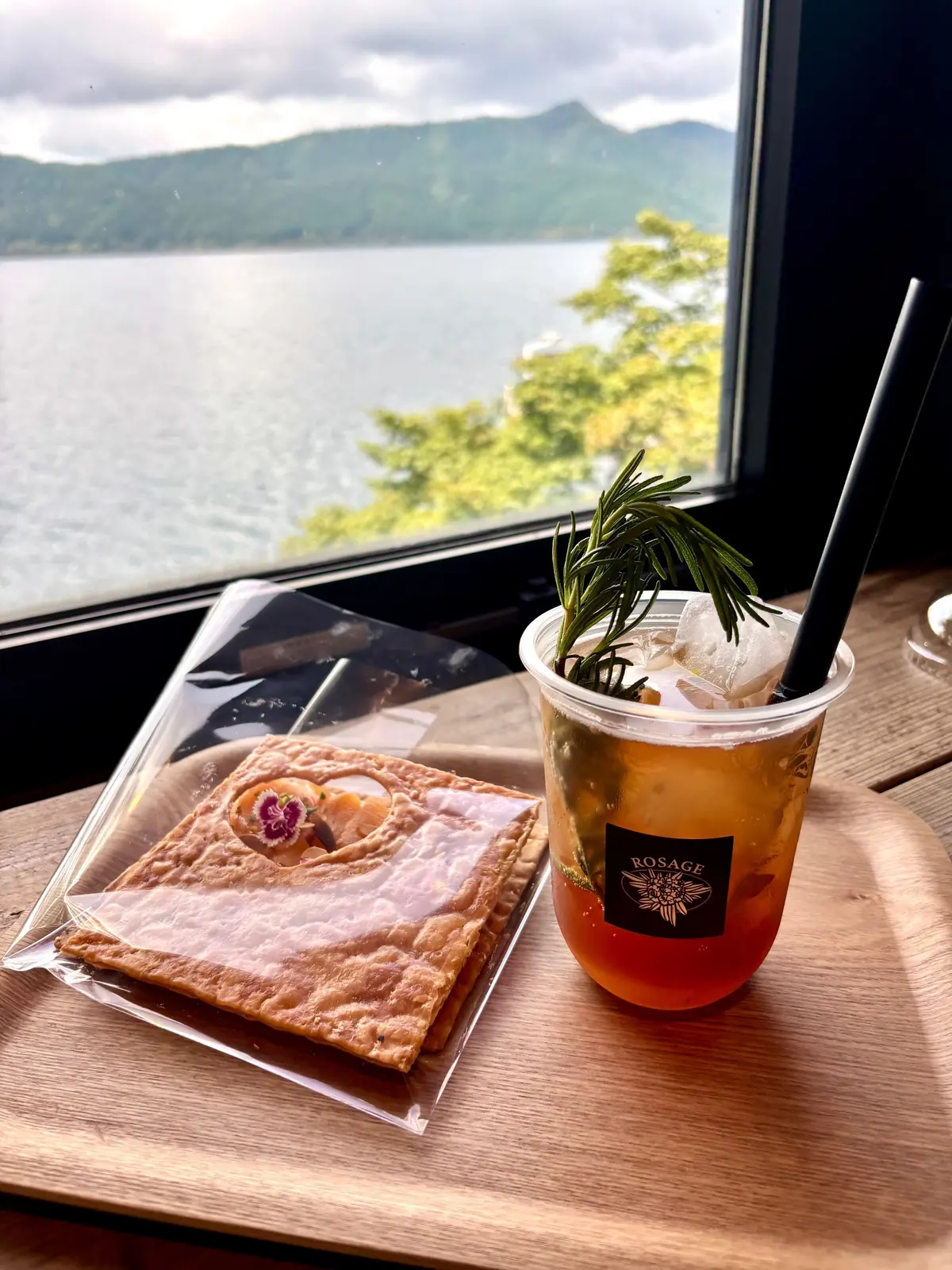 窓から湖の景色を望む木製のテーブルに、ROSAGEのドリンクと焼き菓子が置かれている。ドリンクは氷とローズマリーの葉が入った透明なカップ、焼き菓子は透明な袋に入っている。