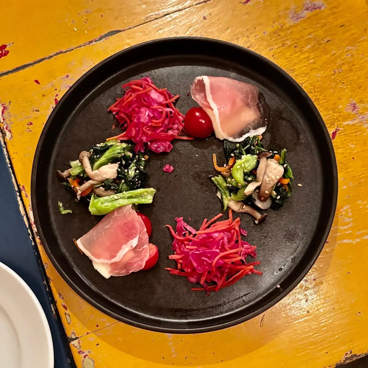 黒い皿に、プロsciutto、即席のキャベツの酢漬け、トマト、きのこ、ほうれん草の炒め物が配置された料理。円形に盛り付けられ、彩り豊か。