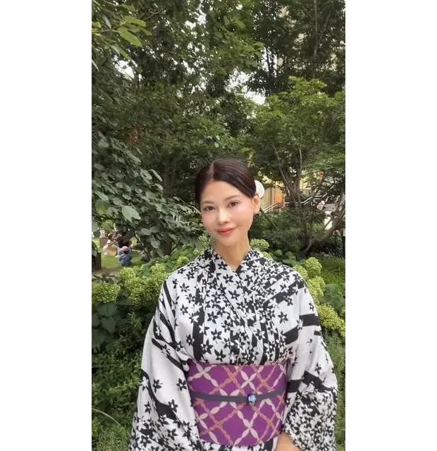白と黒の柄の着物と紫色の帯を身につけた女性。背景には緑豊かな木々が見える。
