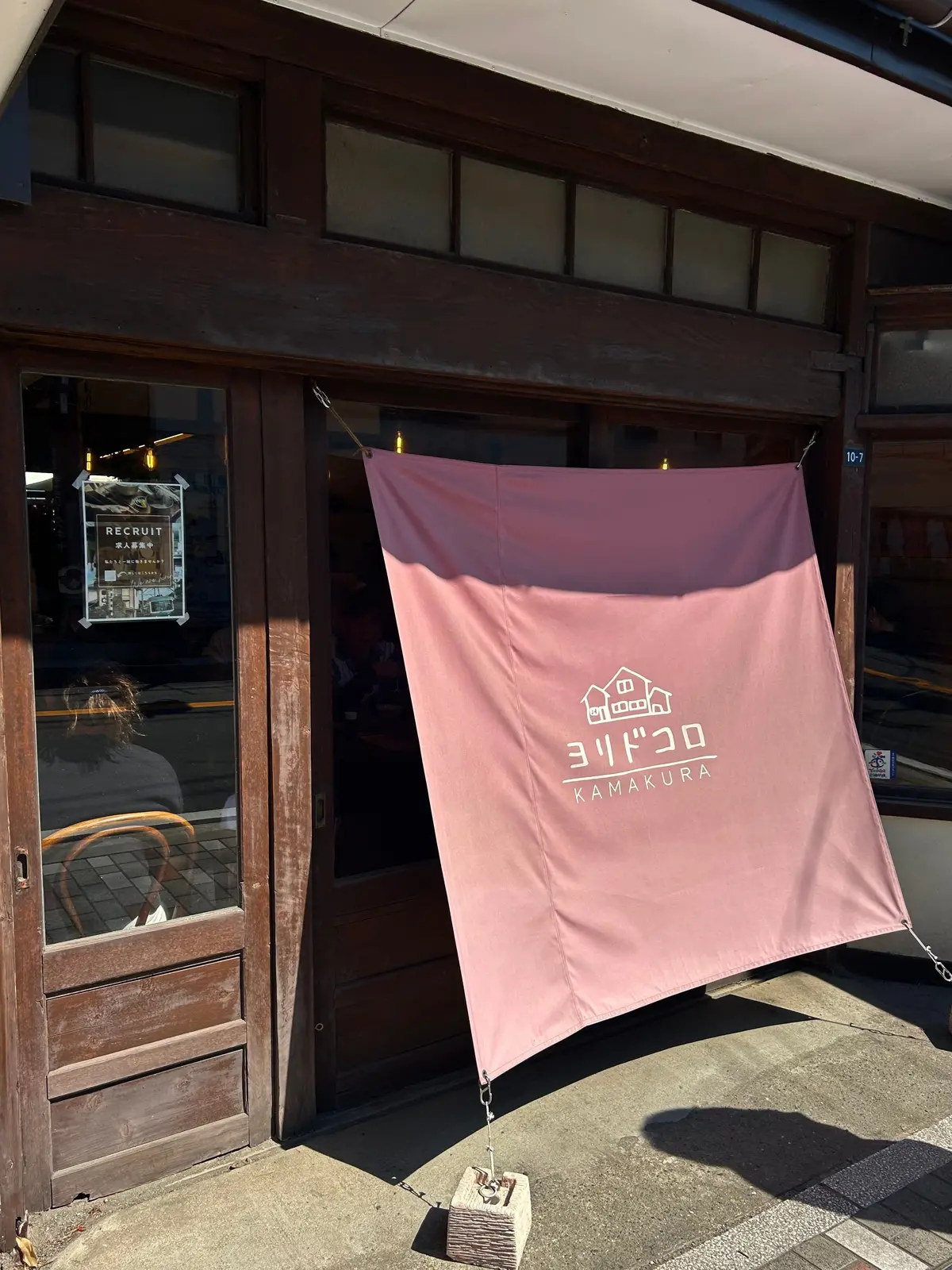 木製の戸口に設置されたピンク色ののれん。のれん中央に白抜きの家型のイラストと「ヨリドコロ KAMAKURA」の文字が配置されている。戸口の窓には「RECRUIT」と書かれた案内板が貼られている。