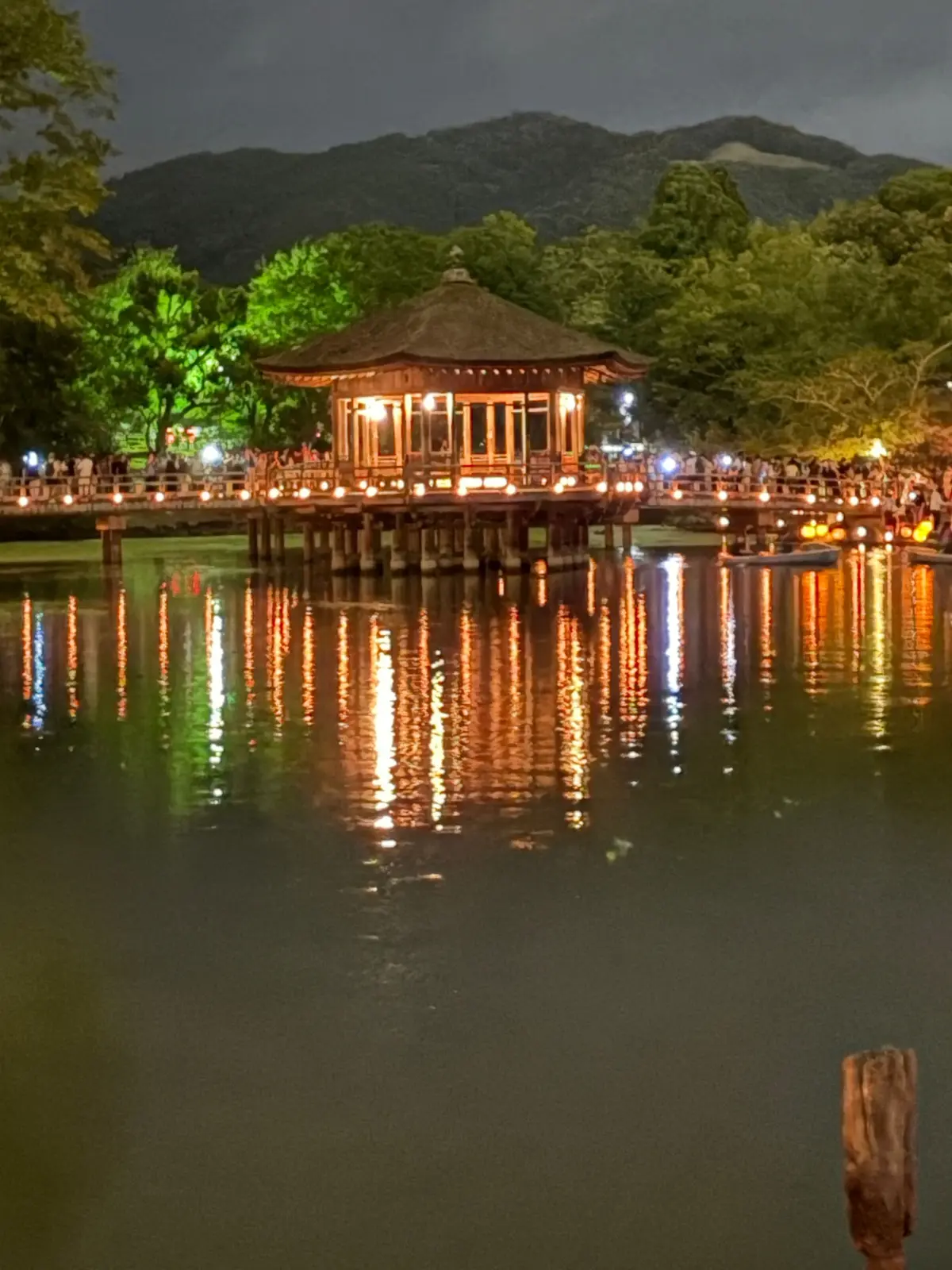 夜の湖に浮かぶ、木造の建物をライトアップした写真。建物は柱で水中に支えられ、周囲に人々が集まっている。水面には光が反射し、幻想的な雰囲気を醸し出している。背景には山が見える。