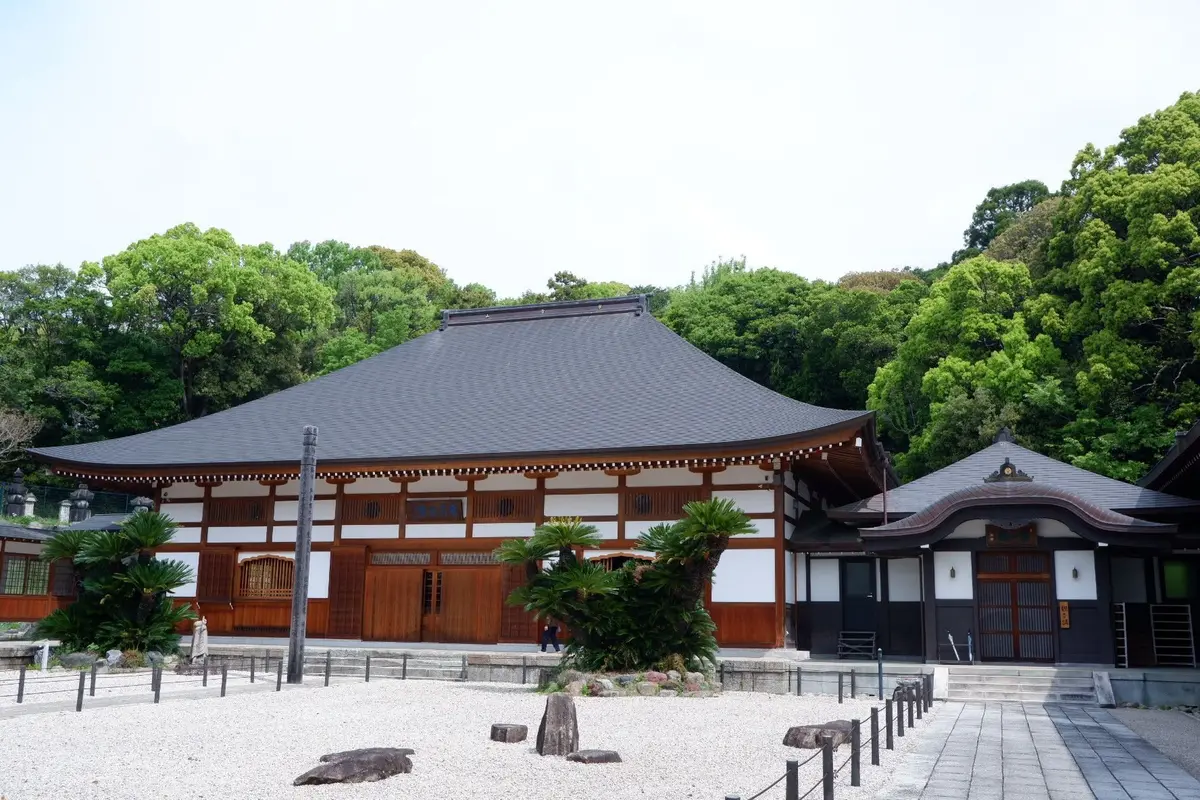 伝統的な和風建築の建物が、緑豊かな木々の中で佇む。建物は黒い屋根と白い壁で、木製の扉と窓が特徴。手前には砂利敷きの空間と岩が置かれている。