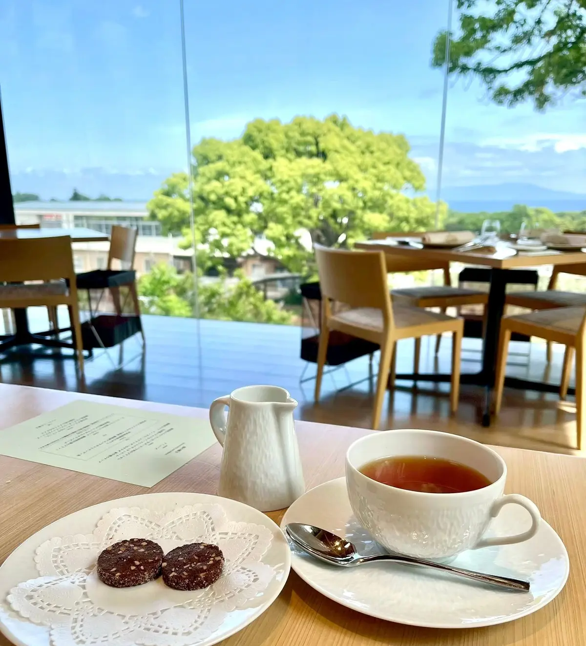 木のテーブルに白いカップと茶色いクッキー、メニューが置かれたカフェの窓から見える緑豊かな景色