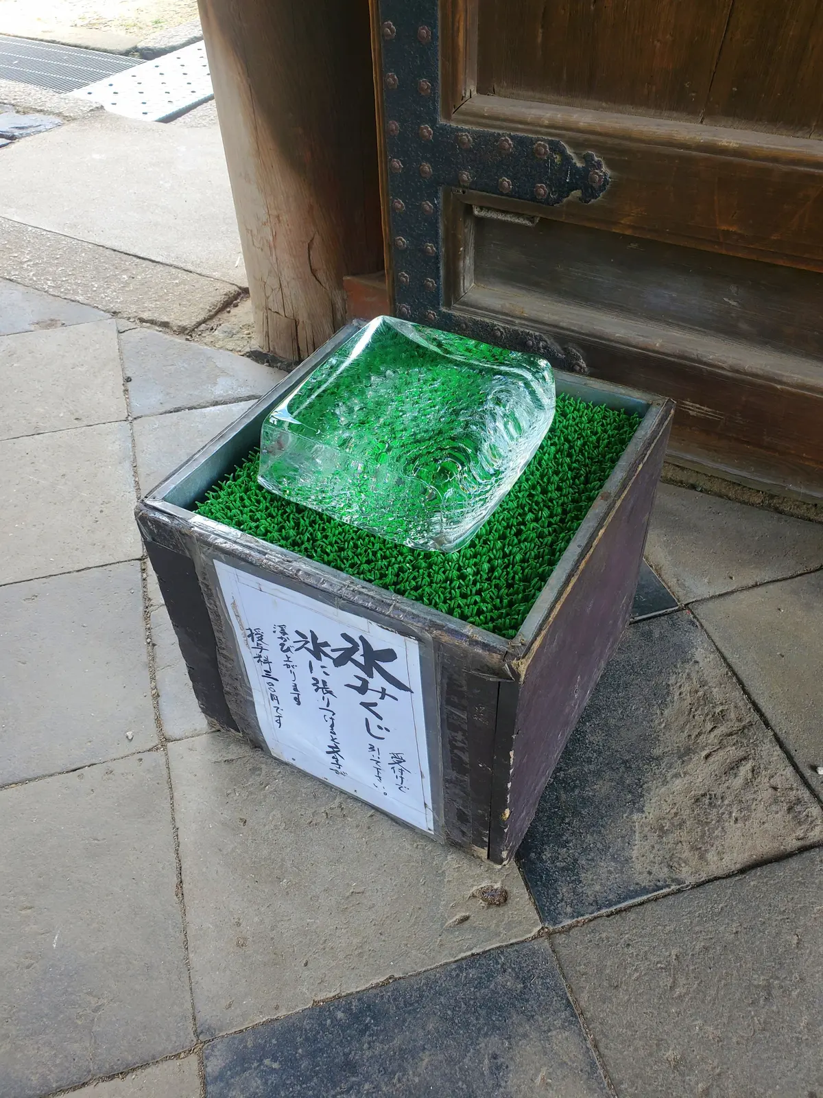 木製の箱の中に、緑の人工芝が敷かれ、透明なガラスの彫刻が置かれている。ガラスは不規則な形をしており、内部に緑色の模様が見える。箱の側面には日本語の文字が書かれている。
