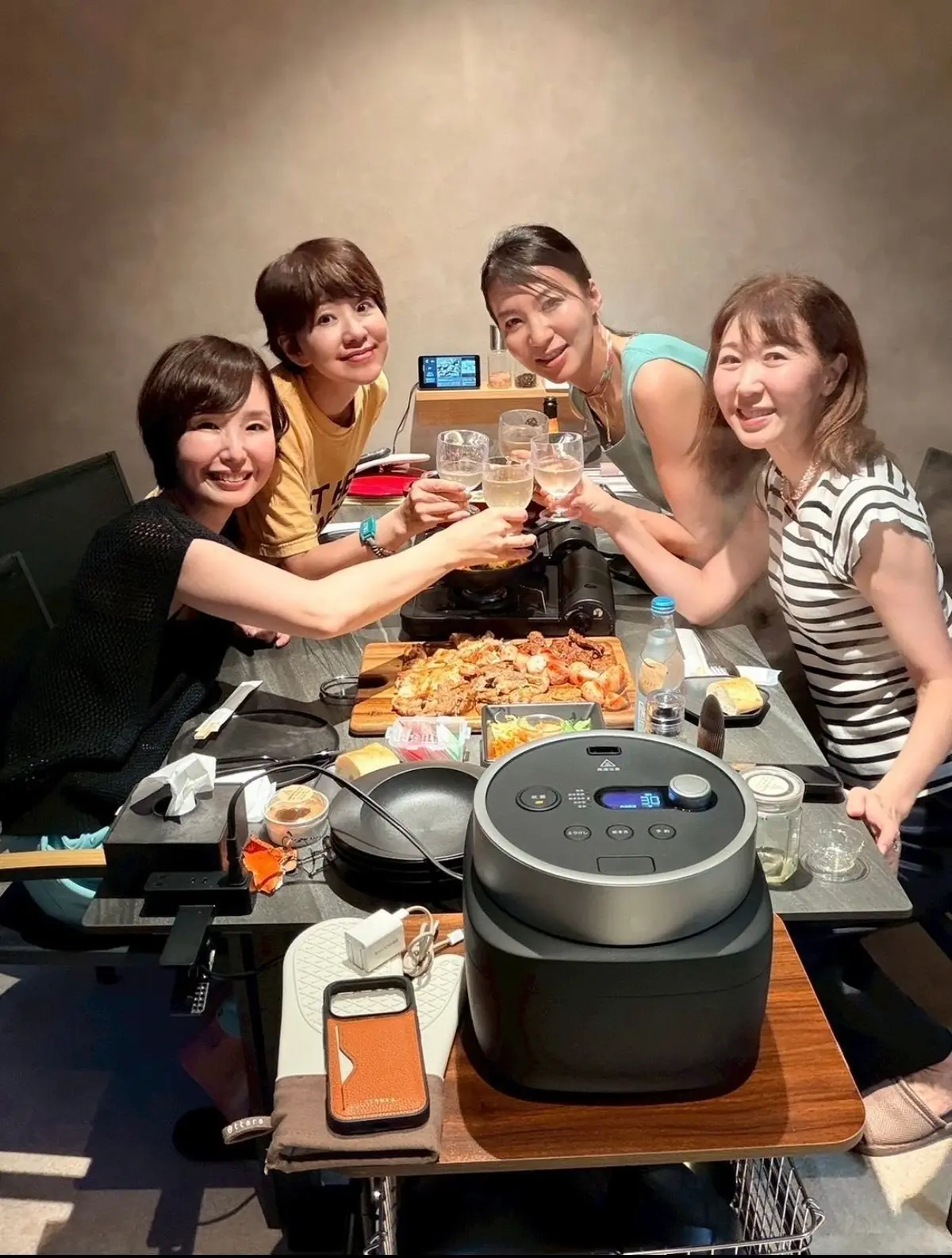 4人の女性がテーブルに座り、グラスを掲げて乾杯している。テーブルには焼き鳥やサラダ、電子レンジやスマートフォンなどが置かれている。