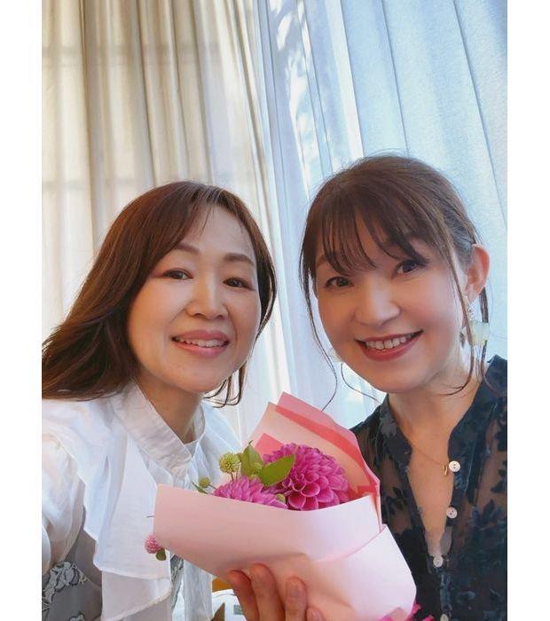 笑顔の女性2人が、ピンク色の花束を持って写っているセルフィー写真。背景には白いカーテンがかかっている。