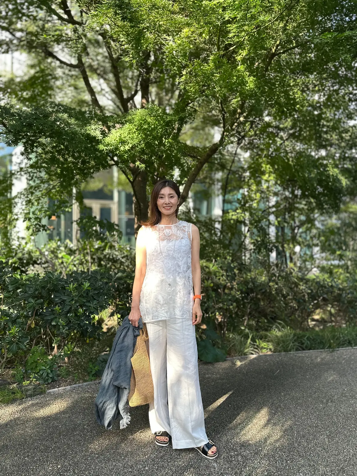 白いパンツと白いレースのトップスを着た女性が、緑豊かな公園で立っている。左手にグレーのショール、右手に編み物のバッグを持つ。背景には建物と木々が見える。