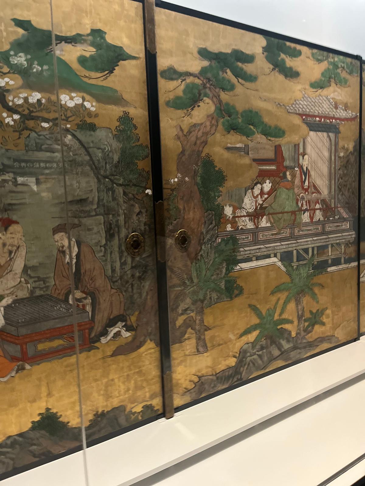 金箔を施した木製の戸棚。左には碁をする老人の姿、右には楼閣と庭園の風景が描かれている。全体的に淡い金色を基調とし、繊細な絵付けが施されている。