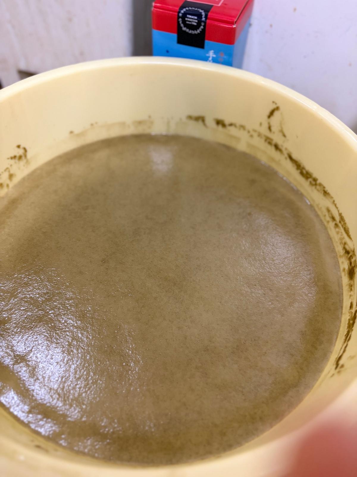 ベージュ色の容器に、表面が平らで均一な濃茶が注がれている。背景には赤と青の箱が少し見える。