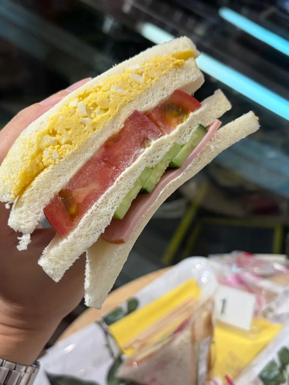 手で保持された卵サンドイッチ。白パンで卵、ハム、トマト、きゅうりが挟まれており、断面から具材が見える。背景には他のサンドイッチが並んでいる。