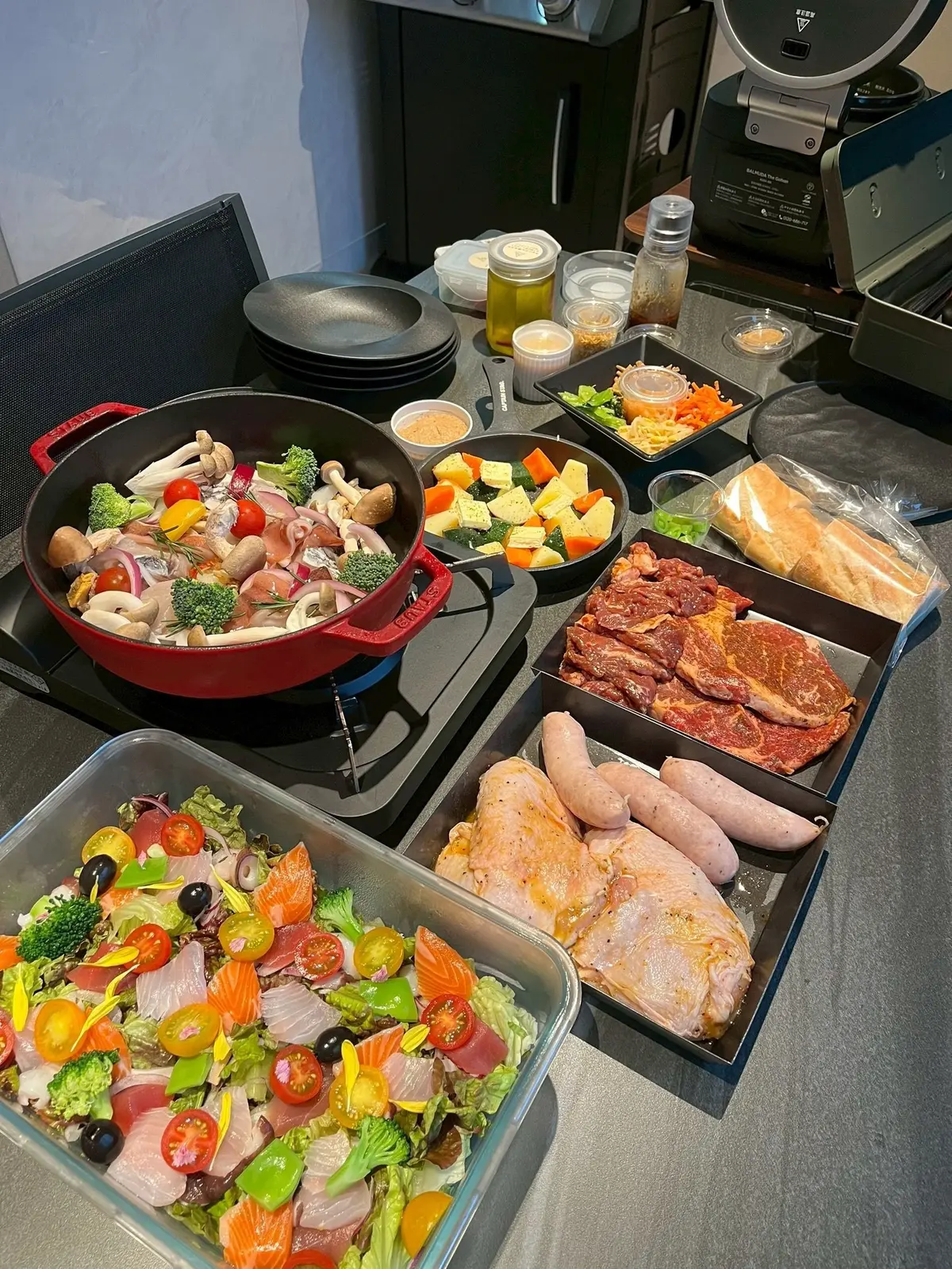 キッチンカウンターに調理済み料理と食材が並ぶ。赤のホットプレートには野菜と肉、ガラス容器には色とりどりのサラダ、トレイにはステーキ、ソーセージ、鶏肉が置かれている。