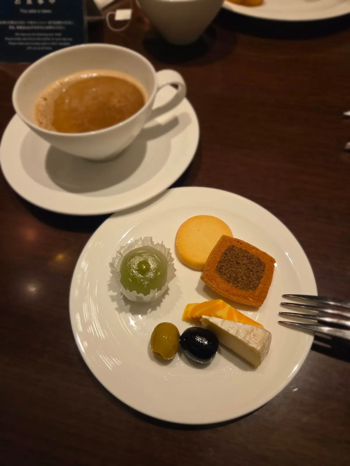 コーヒーのカップとソーサー、それに様々な軽食が盛り付けられた白い皿が写っている。皿には、緑色のゼリー、クッキー、ハーブ風味のケーキ、オリーブ、チーズ、柑橘系のスライスなどが配置されている。