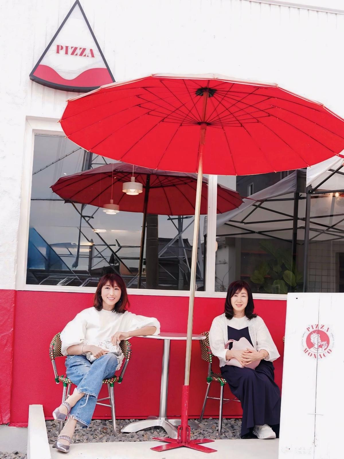 白い壁のピザ屋の前に、赤い傘の下で2人の女性が座っている。左の女性は白いトップスにデニム、右の女性は黒いトップスに紺色のパンツを着用。背景にはピザの看板と、金属製のパーゴラが見える。