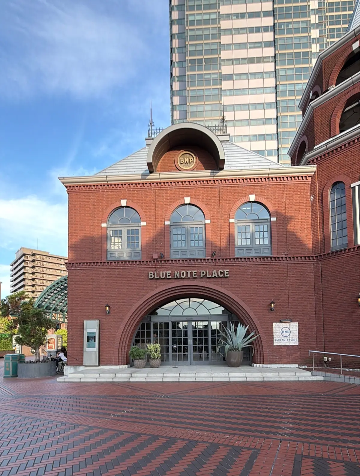 レンガ造りの「BLUE NOTE PLACE」入り口、中央にアーチ型のガラスドア、左右に植えられた観葉植物、背景に高層ビルが立つ。