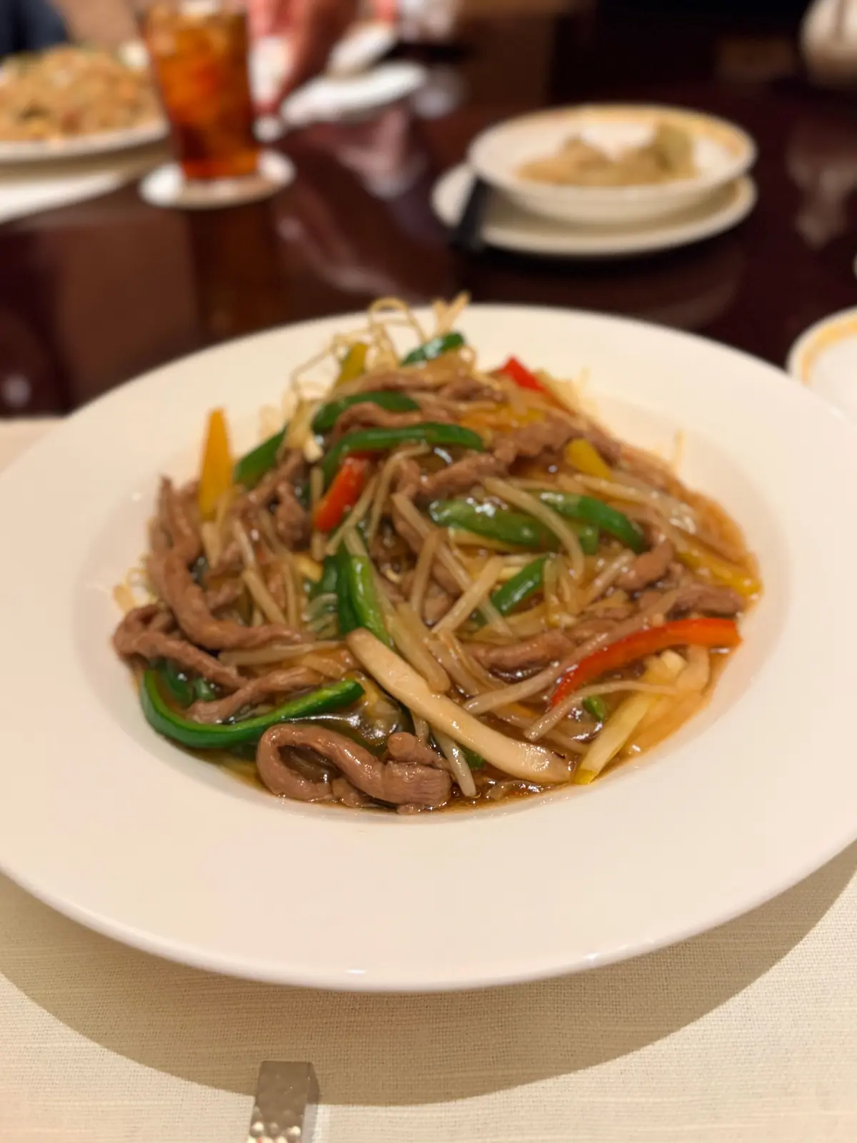 白い皿に、牛肉と野菜の炒め物が盛り付けてある。緑のピーマン、赤のパプリカ、細い麺、きのこが混ざり、茶色のソースが全体を覆っている。