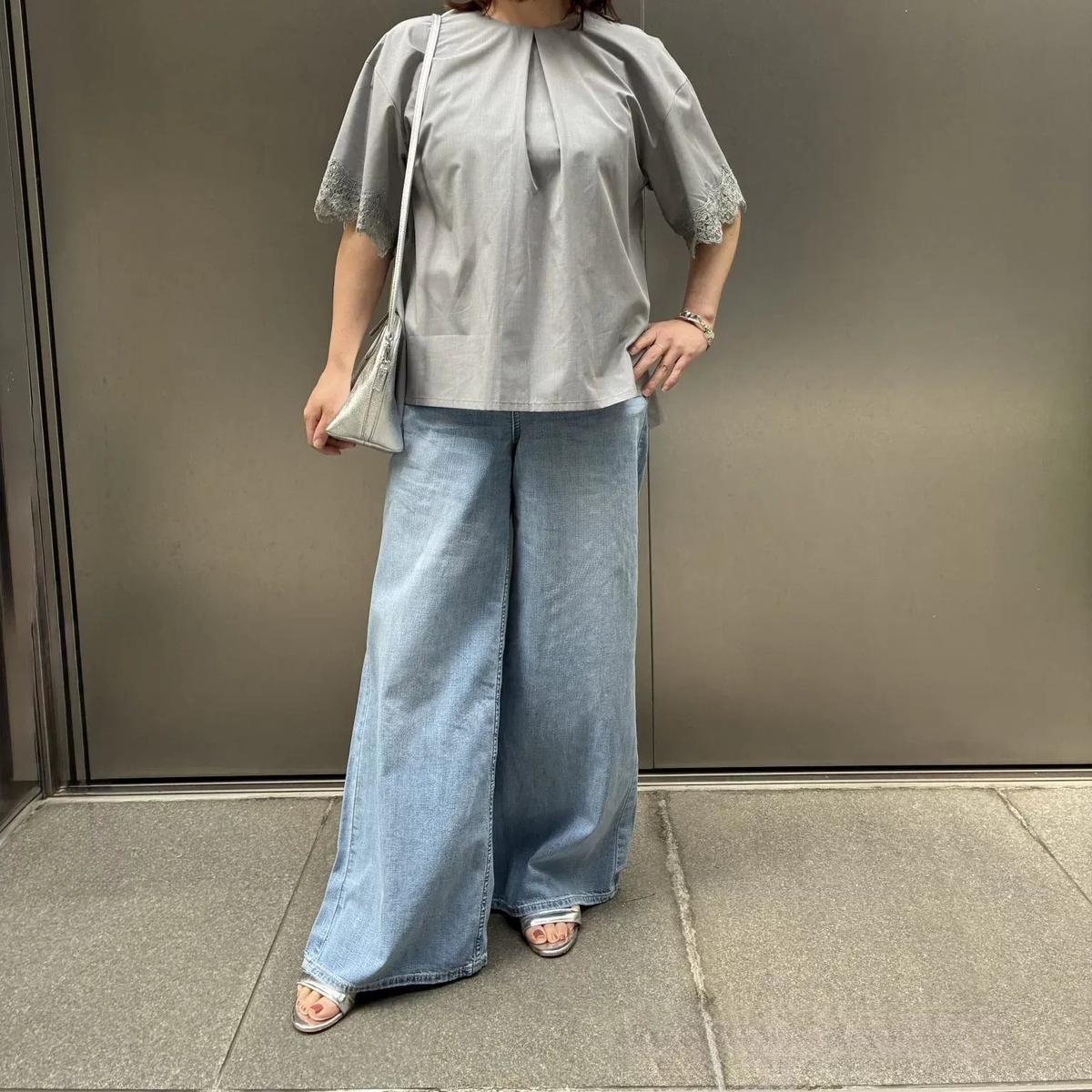 シルバーのリボンが付いたグレーのトップスと、ゆったりしたデニムパンツを着た女性が、グレーの壁の前でポーズしています。左手には小さめのシルバーのハンドバッグを下げ、右手はウエストに置いています。足元にはベージュのサンダルを履いています。