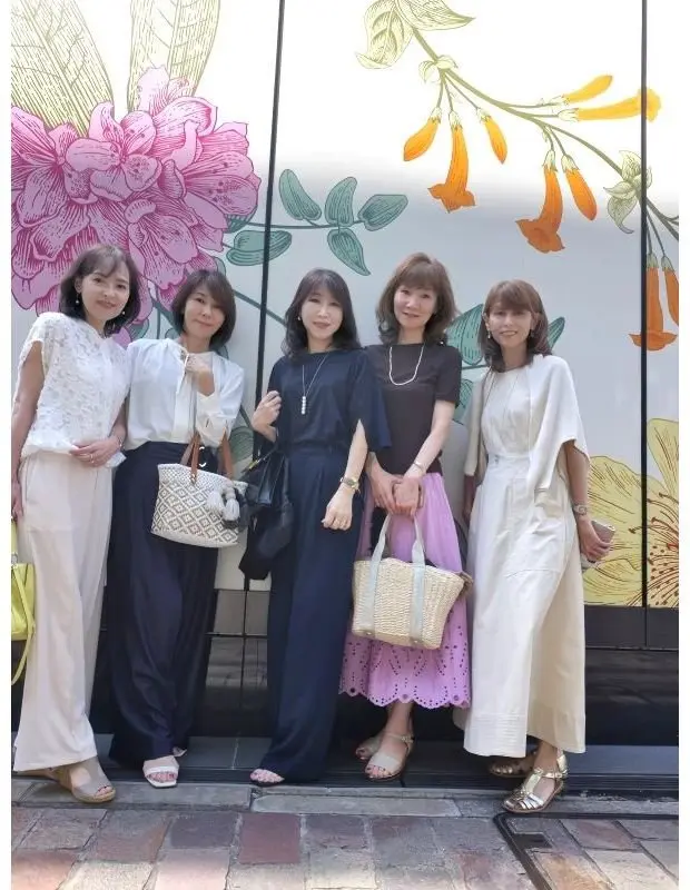 5人の女性が並んで立ち、花の模様が描かれた壁の前でポーズをとっている。左から白のトップスと白いズボン、白のブラウスとネイビーのズボン、ネイビーのワンピース、黒のトップスとピンクのスカート、白のワンピースを着用。各自がカバンやアクセサリーを持っている。