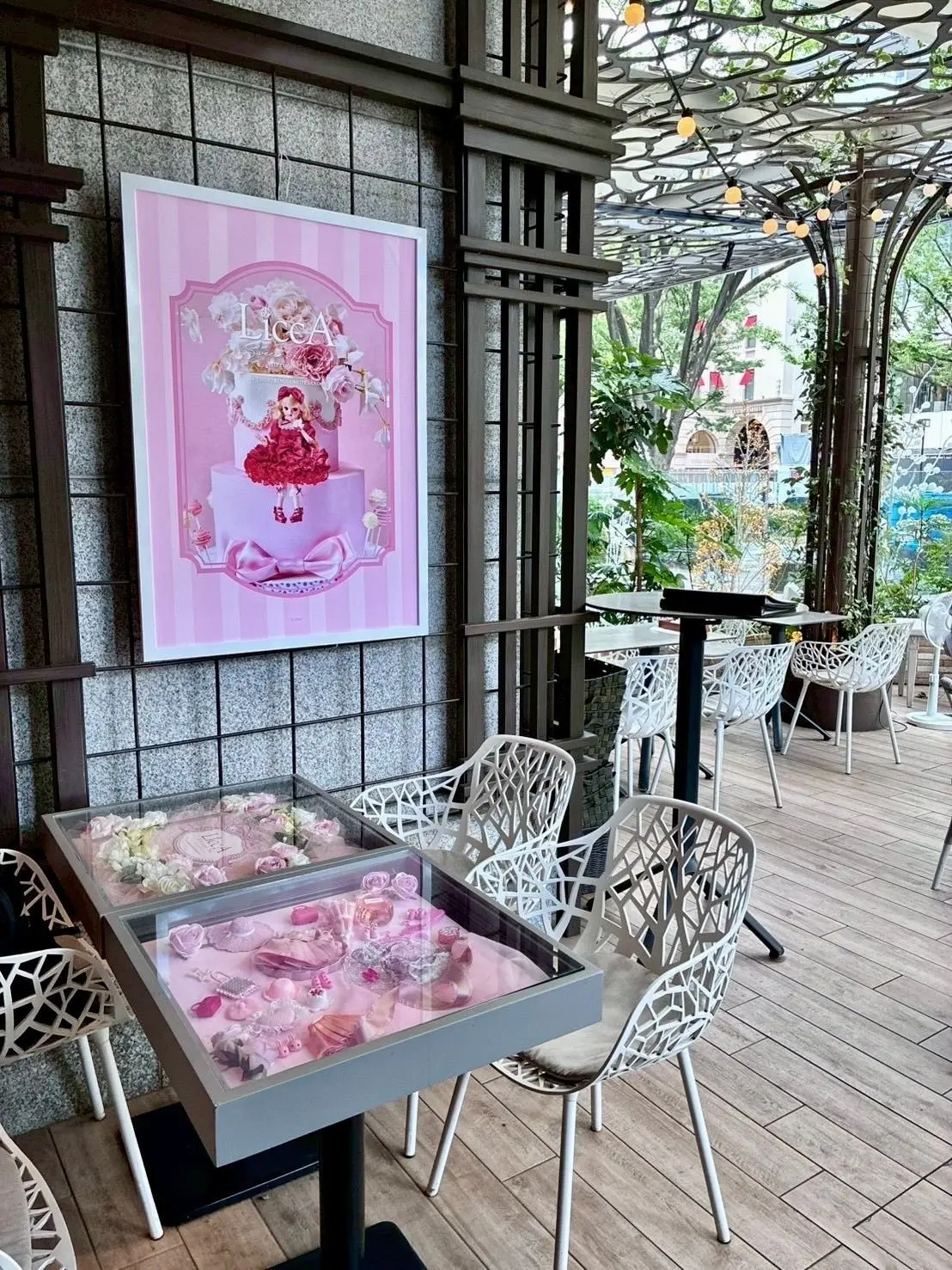 ゼフコヴァのテラス席にあるリカちゃんコラボのガラステーブル席。壁に飾られた額に入った、ピンクと白のストライプ模様のデザイン。中央に「Licca」という文字と花が飾られ、ピンクのドレスを着た人形が二段のケーキの上に立っている。ケーキにはリボンや小さな装飾が施されている。