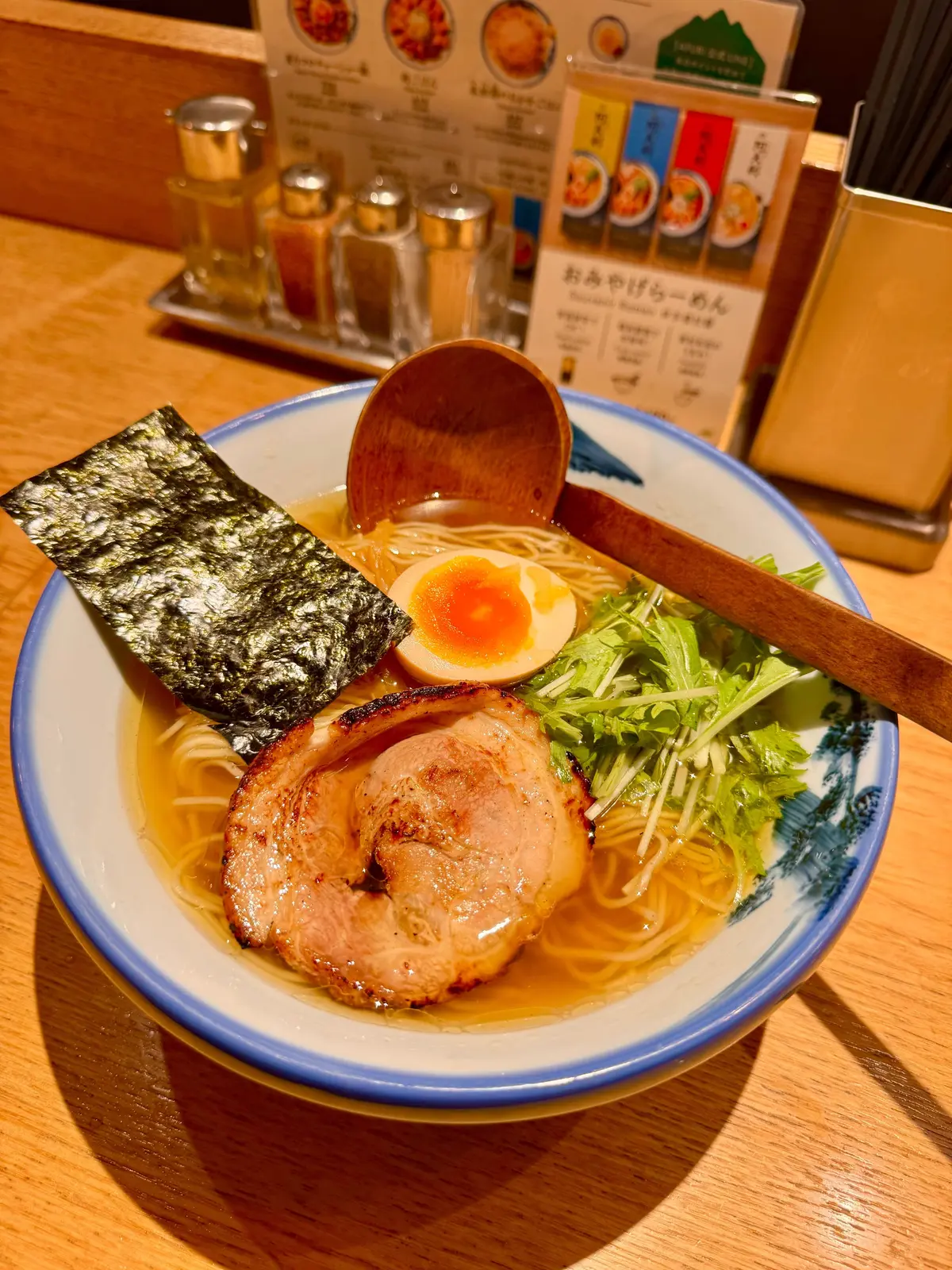 青い丼に入ったラーメン。豚肉、海苔、ネギ、半熟卵が乗っている。木のレンゲが添えられ、背景にはメニューが置かれている。