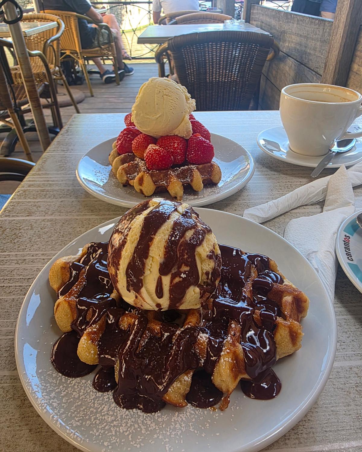 テーブルに置かれた、ワッフルとフルーツのデザート、コーヒー。手前はチョコレートソースをかけたワッフルにアイスクリーム、奥はフルーツとワッフル、さらにアイスクリームが乗っている。白い磁器の皿に盛り付けられ、背景にはカフェのテーブル席が見える。