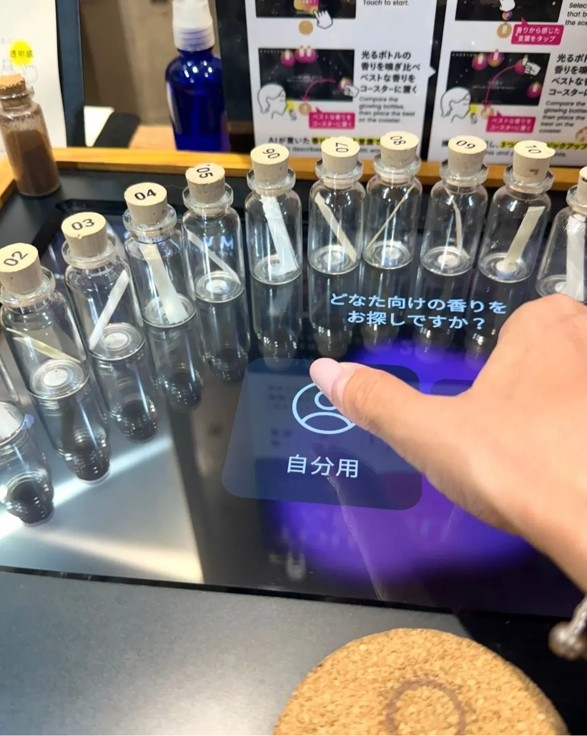 透明なガラス瓶が横一列に並び、それぞれに番号が貼られている。前面にタッチパネル式のモニターがあり、「自分用」のボタンが光っている。背景には説明書きが見える。