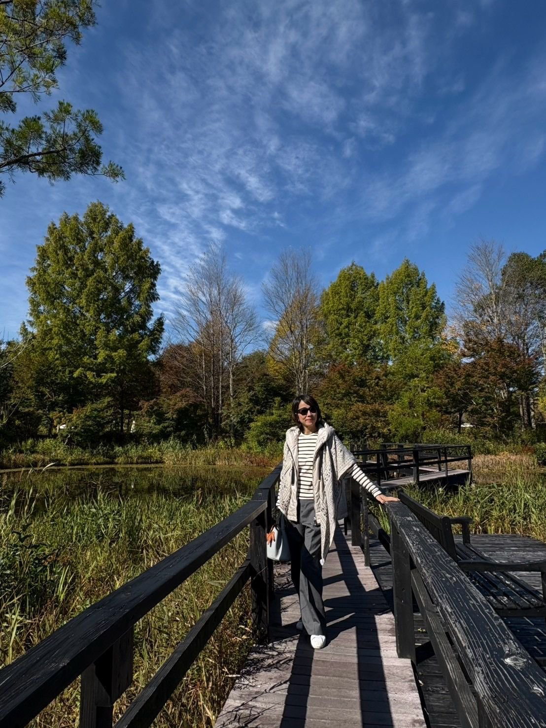 木橋の上を歩く女性。白いボーダーのトップスにベージュのパンツ、白いカーディガンを羽織っている。背景には水辺の緑豊かな植生と、青空。