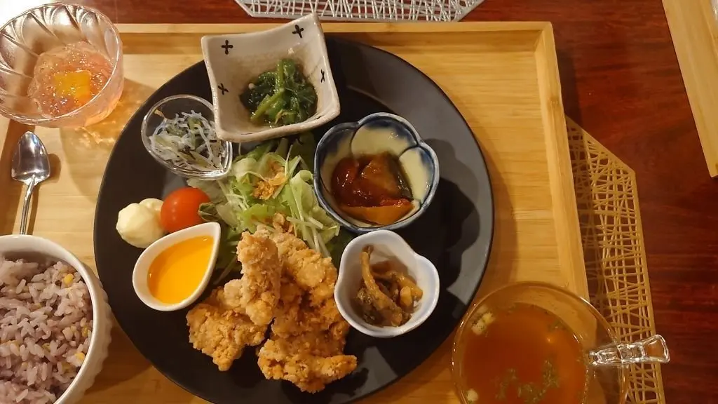 木製のトレイに盛り付けられた和食の定食。黒い皿に、鶏の唐揚げ、卵焼き、サラダ、お味噌汁、煮物、漬物などが彩り豊かに配置されている。白米が別添えの小鉢に入り、飲み物と思われる透明な液体とオレンジ色のスープも添えられている。