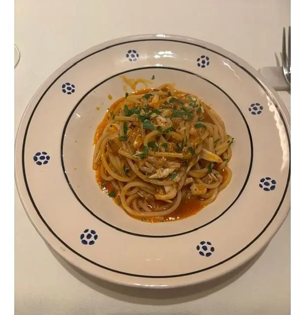 白地に青い模様の皿に盛り付けられた、パスタ料理。細めの麺はオレンジ色のソースでコーティングされ、白い具材と緑のハーブが散らされている。
