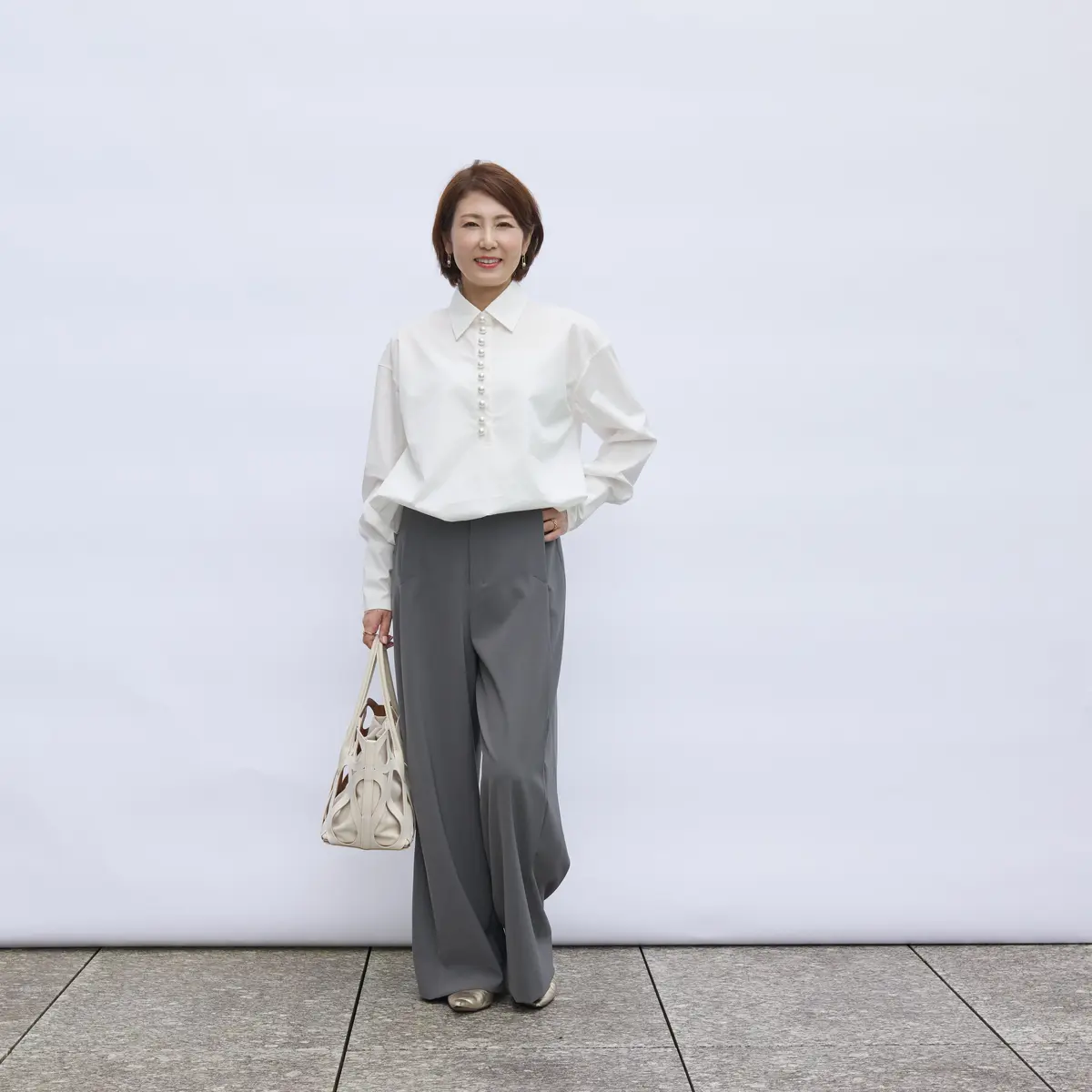 白いボタンダウンシャツにグレーのワイドパンツを合わせた女性。右手にベージュのハンドバッグを持ち、上品な装い。背景は白い壁とグレーのタイル張りの床。