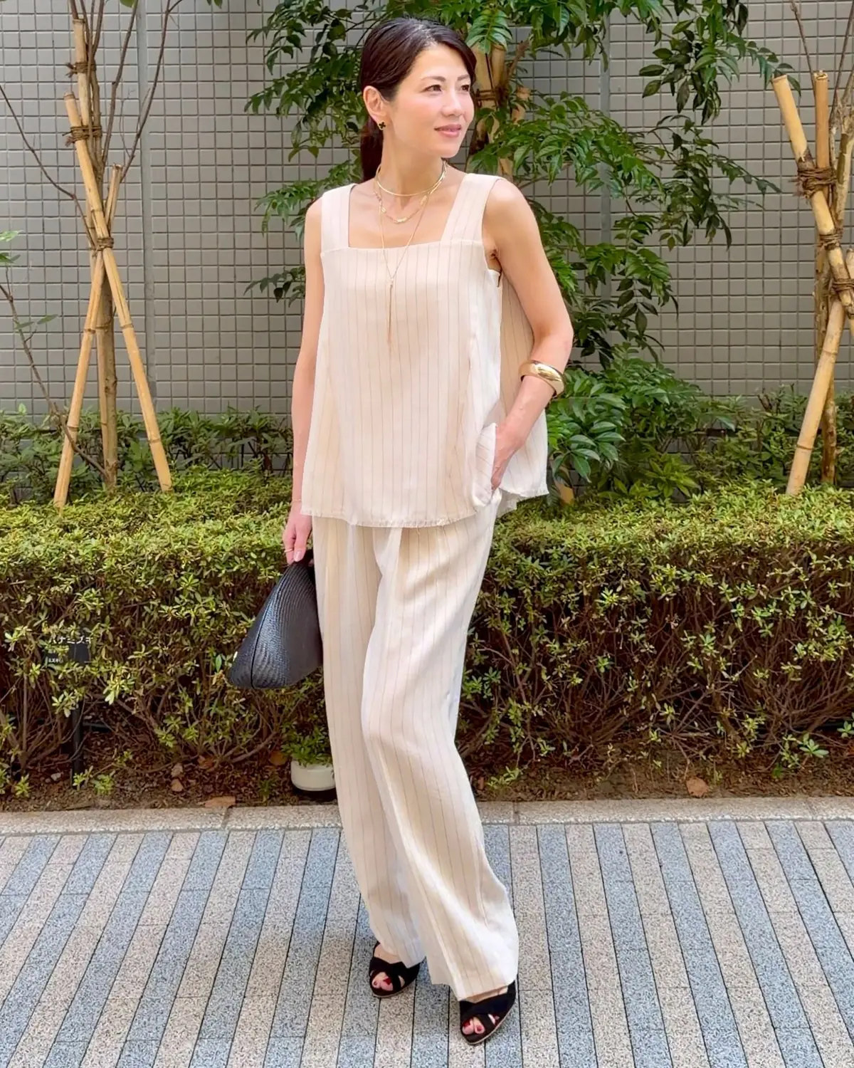 ベージュのパンツとトップスを着用した女性が立っている。黒いサンダルを履き、複数のネックレスを身につけている。黒い扇形のバッグを持ち、背景には緑の植物がある。
