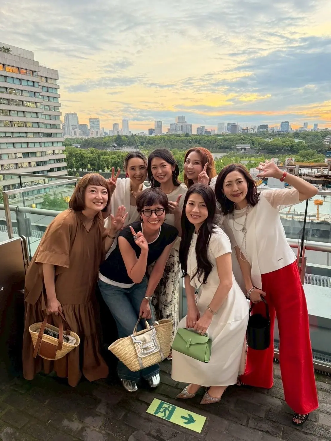 屋上から、7人の女性が夕焼けを背景に笑顔で写っている。様々な色の服を着ており、手でヴィクトリーサインをしている人がいる。手前に緑色の矢印の標識が見える。
