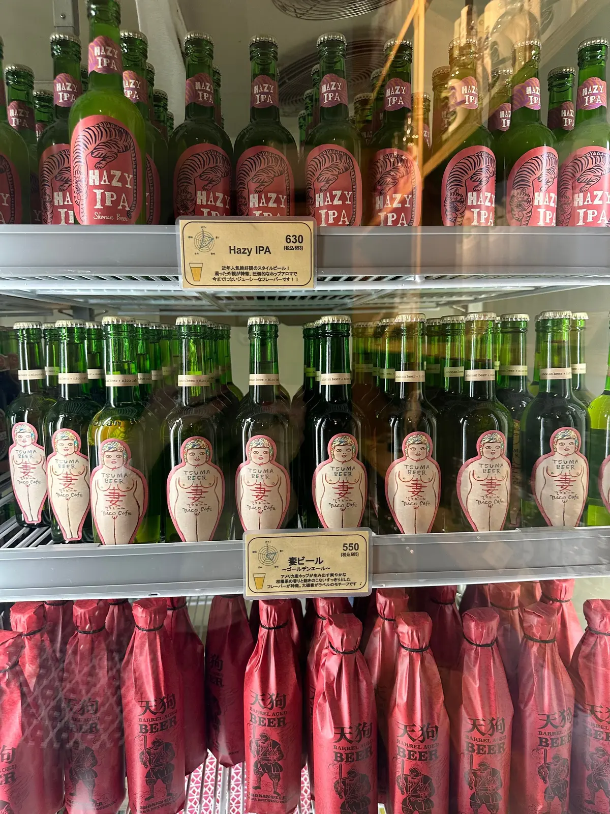 ハジーIPAとツマビールのボトルが並んだ冷蔵棚。ハジーIPAは緑のガラス瓶にピンクのラベル、ツマビールはピンクのボトルに女性像のデザインが施されている。