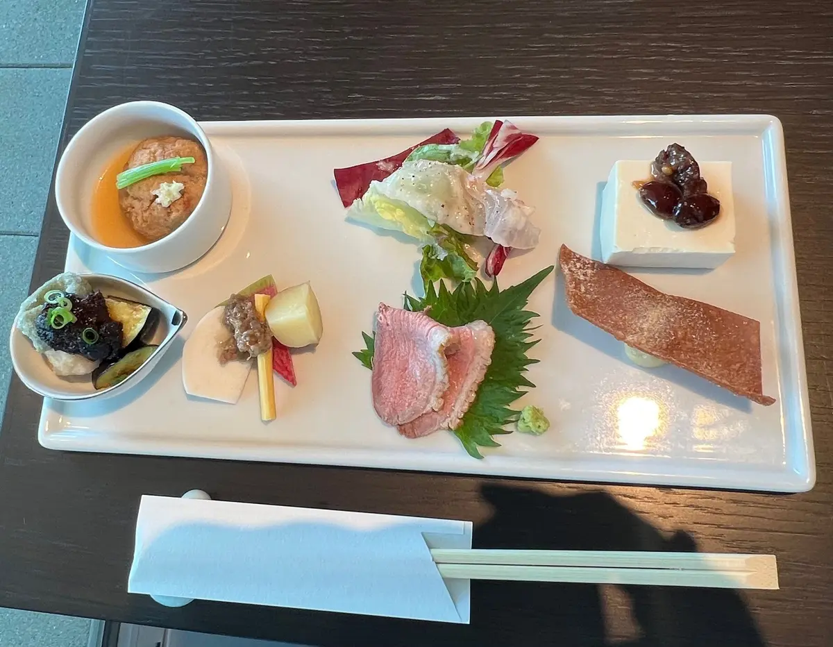白い長方形のプレートに、様々な料理が美しく配置されている。中央にはレタスとラディッシュのサラダがあり、右にはピンク色の肉とワサビの葉、左には小鉢と揚げ物、小さなボウルにスープが置かれている。