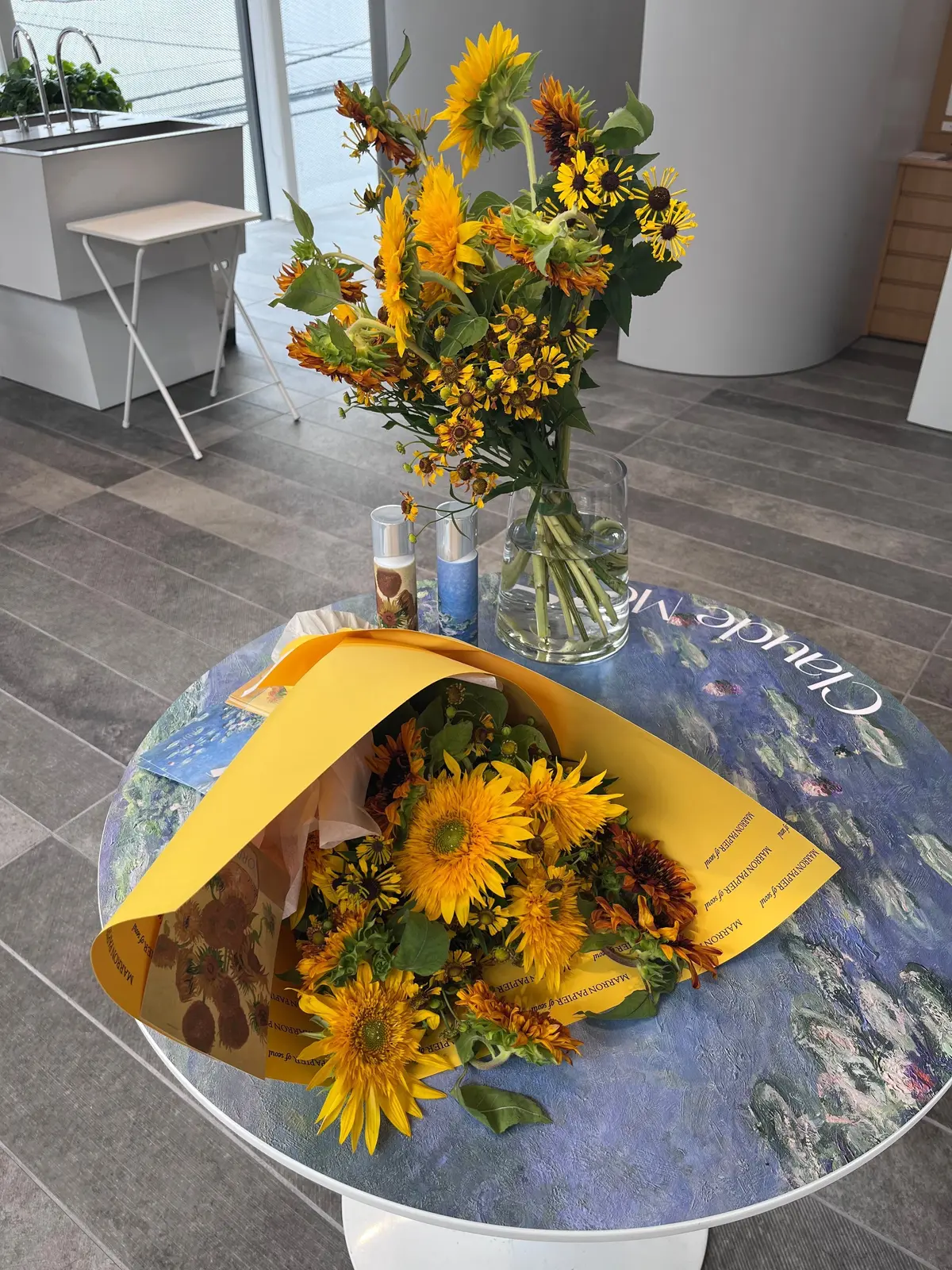 丸いテーブルに、黄色いひまわりやオレンジ色の花束が置かれている。背の高い花束はガラス瓶に活けられ、手渡しの花束は黄色い包装紙で包まれている。背景には、タイル張りの床と窓が見える。