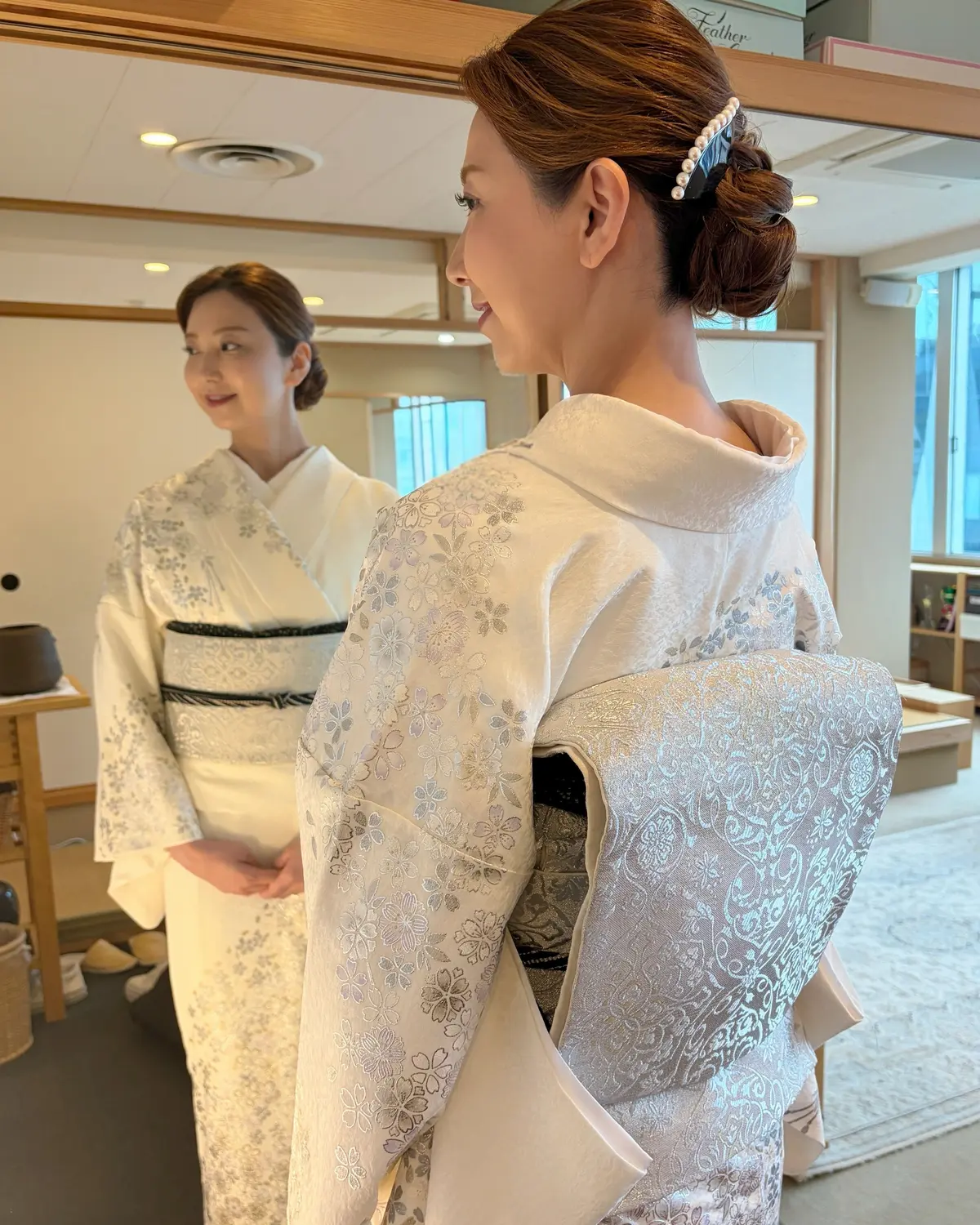 白い花模様の着物を着た女性が、ミラーに映る自分の姿を見つめている。後ろには同じ着物を着た別の女性が立っている。