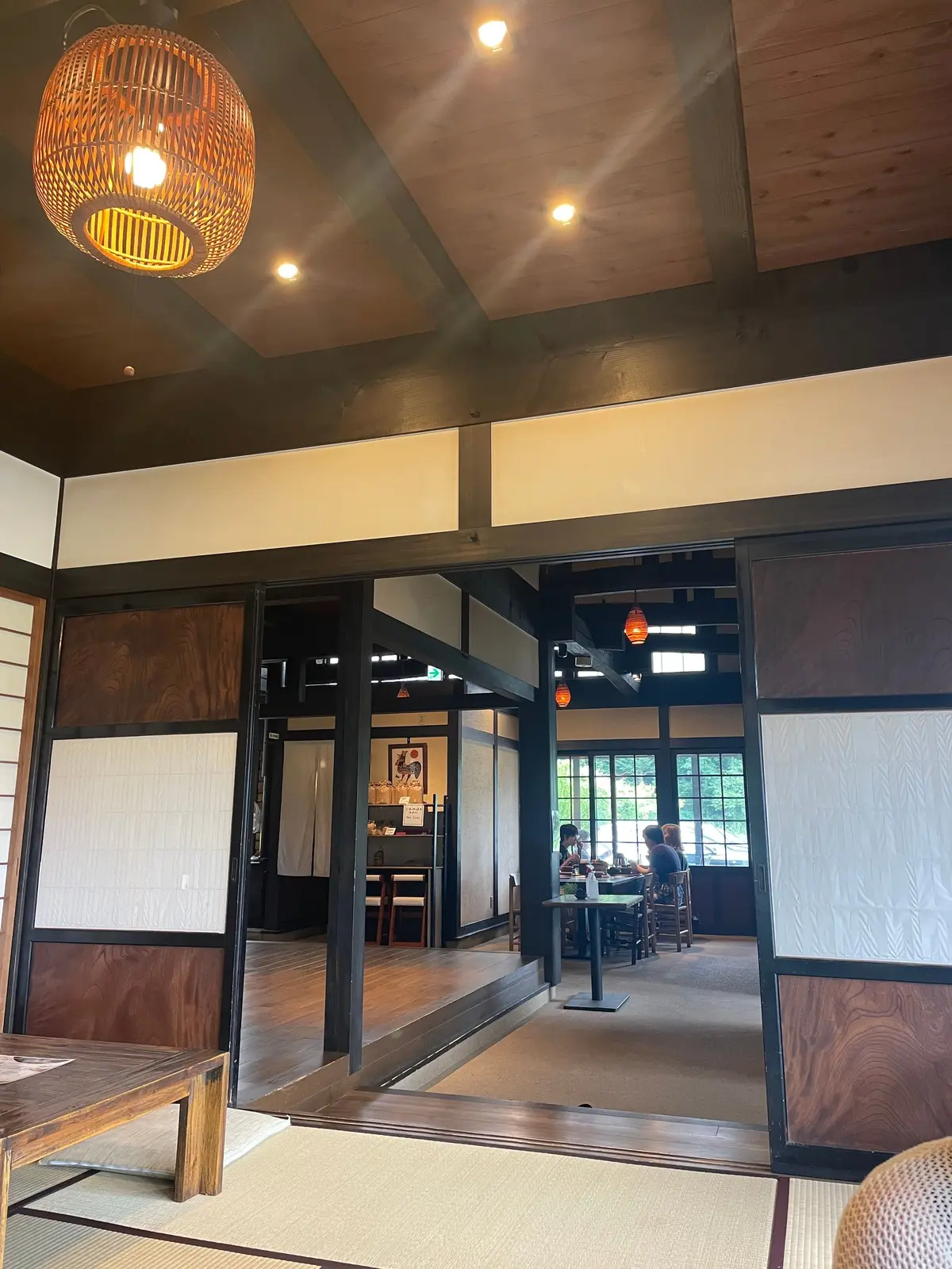 木目調の天井と白い障子で仕切られた和風の空間。手前に編み込まれたデザインの照明器具、奥にはテーブル席と窓が見える。