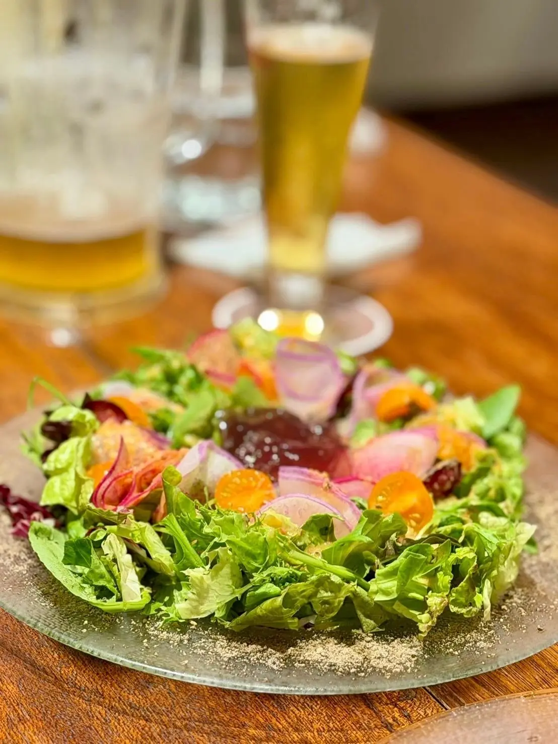 木製のテーブルに置かれた、緑色の葉野菜を中心としたサラダ。赤やオレンジ色の食材、薄紫色の輪切りが彩りを添えている。背景にはビールグラスが写る。