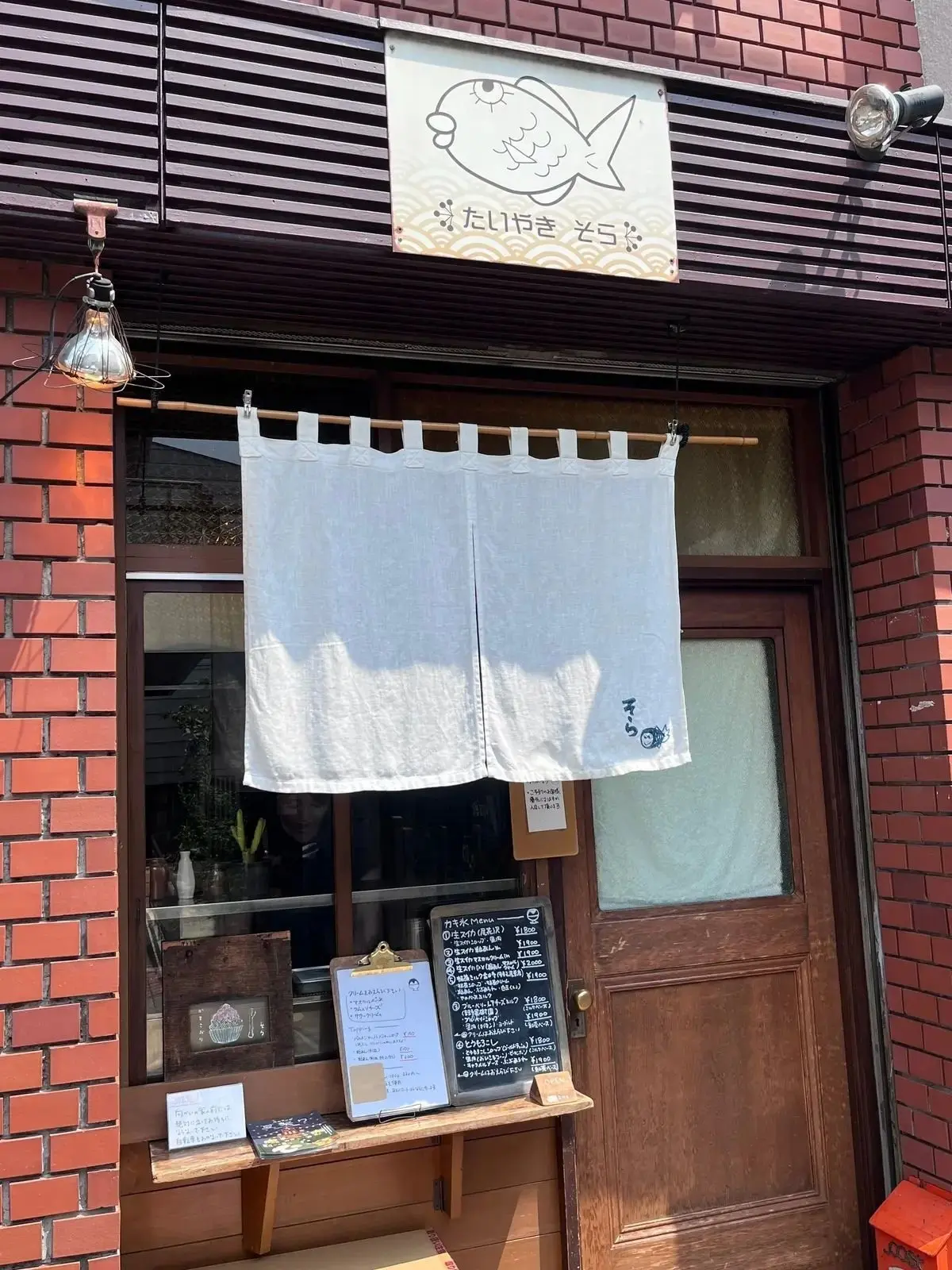 茶色いレンガ張りの外壁に、大きなガラス窓と木製のドアがあるお店の正面。窓には白いカーテンがかかり、店名が書かれた看板が上部に設置されている。看板には、魚のイラストと「たじりやき 乞々」の文字。ドアの横にはメニューが書かれた黒板。