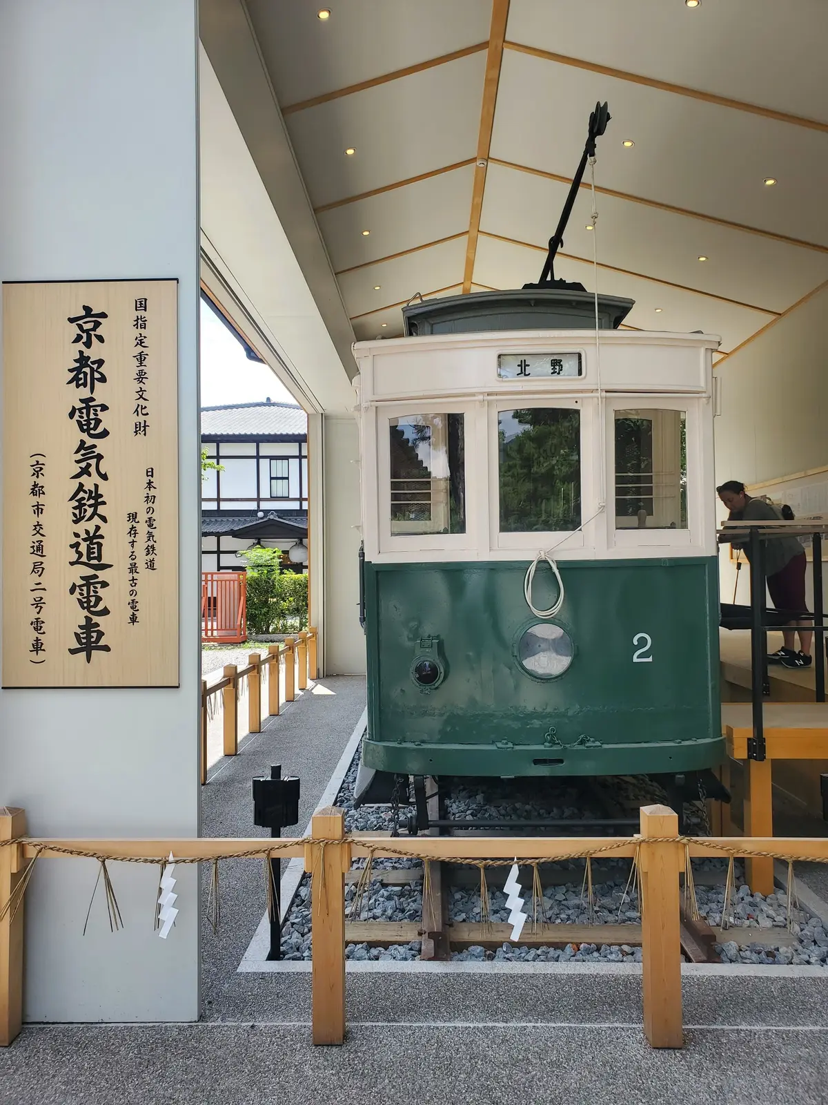 京都の路面電車「京都市電」の保存車両。緑色の車体、前面に大きな窓、屋根には集電装置。展示会場に置かれ、背景には解説の看板が立っている。
