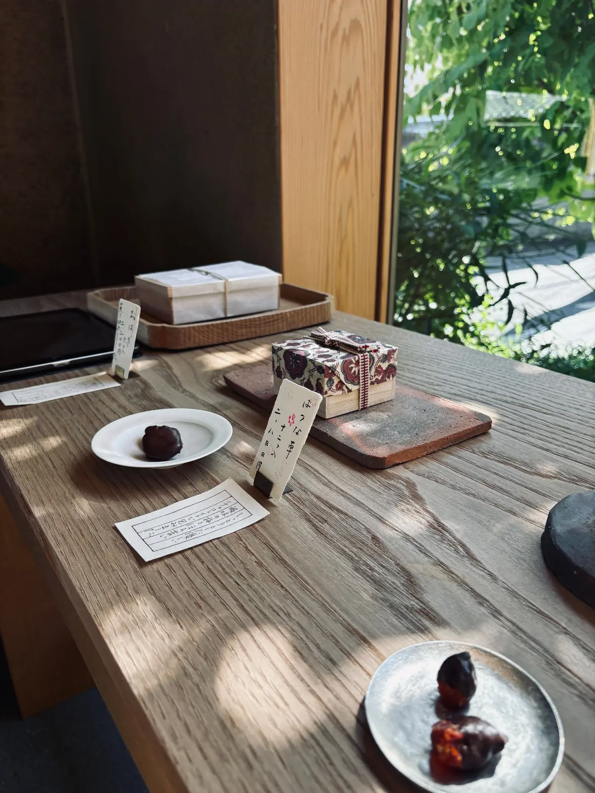 木製のテーブルに、小さな白い皿にチョコレートがのった料理と、花模様の包装されたお菓子が並んでいる。窓の外には緑の木々が見える。