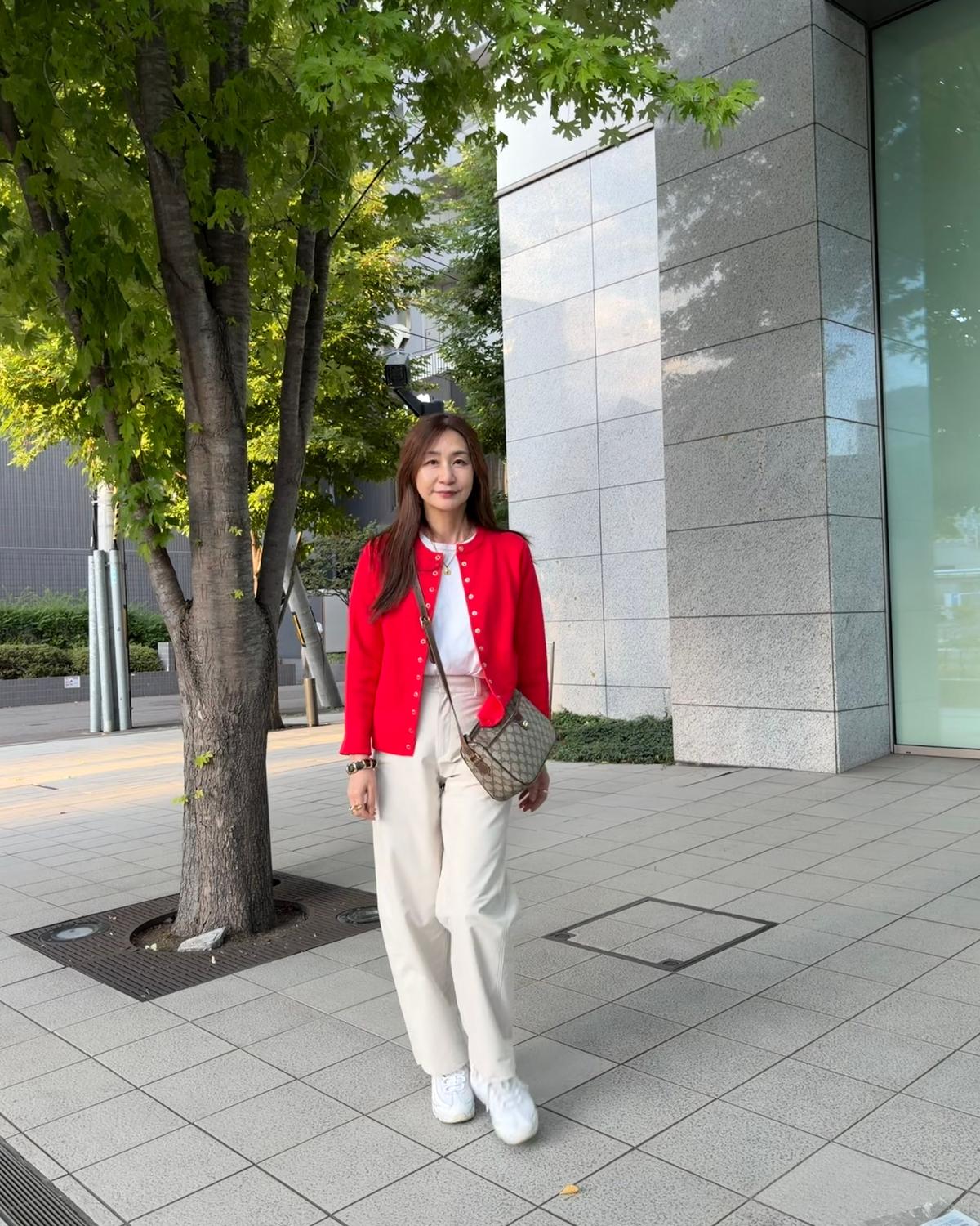 赤いカーディガンに白いパンツ、白いスニーカーを着用した女性が、建物の前で立っている。右手に茶色のバッグを持つ。背景には緑の木とグレーの建物がある。