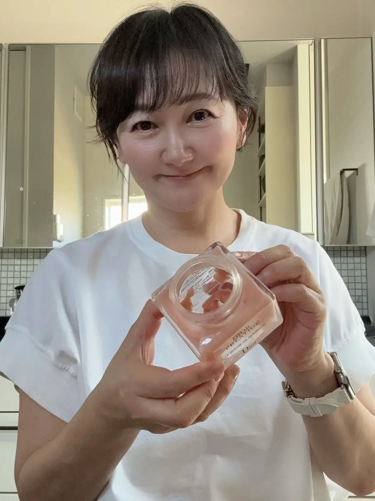 白いTシャツを着た女性が、ディオールのリップグロスを持ち上げて見せている。背景には白い洗面台と鏡、グレーのタイルが映る。