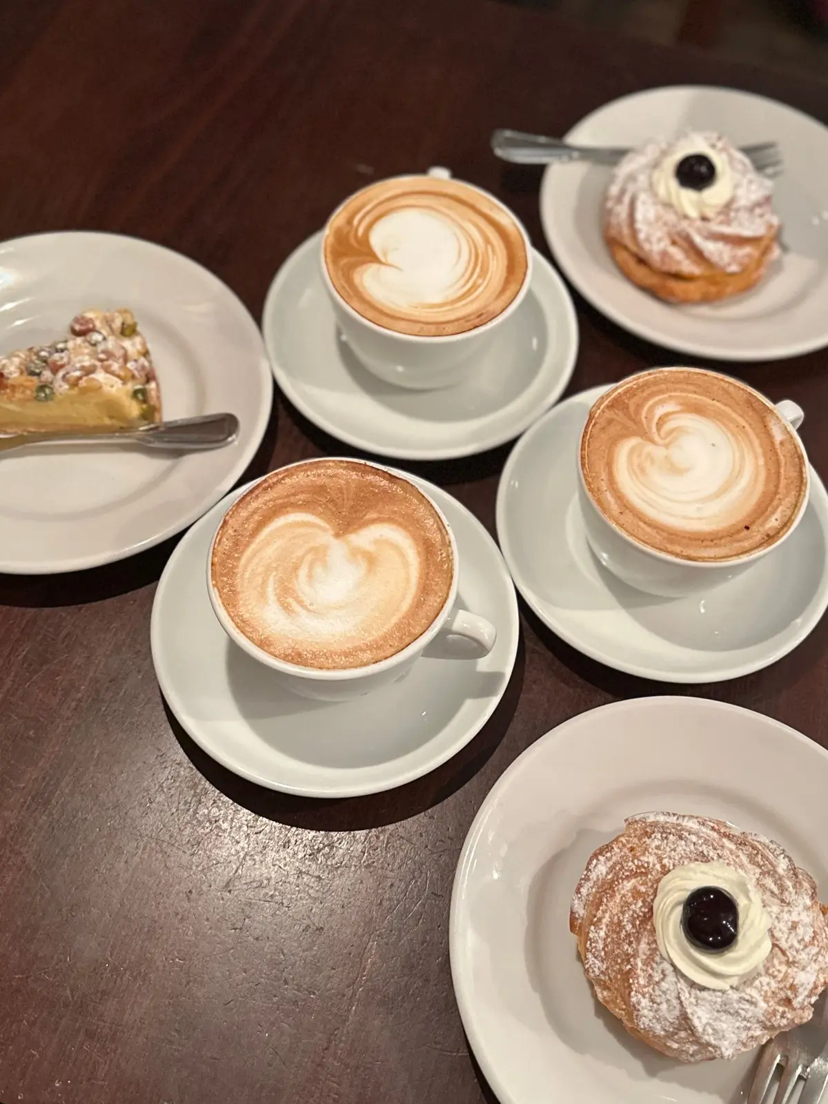 木製のテーブルに置かれた、コーヒーと焼き菓子の盛り合わせ。2つのカフェラテ、ケーキの切れ端、粉糖をかけた焼き菓子が白磁の皿に載っている。