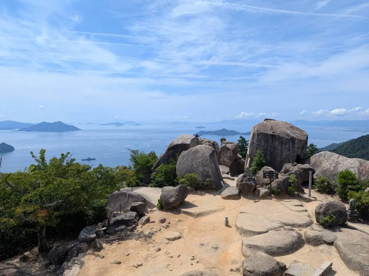 山頂からの眺望。青い空と白い雲、遠くには島々が点在する海。岩場に休憩スペースがあり、人が数人いる。