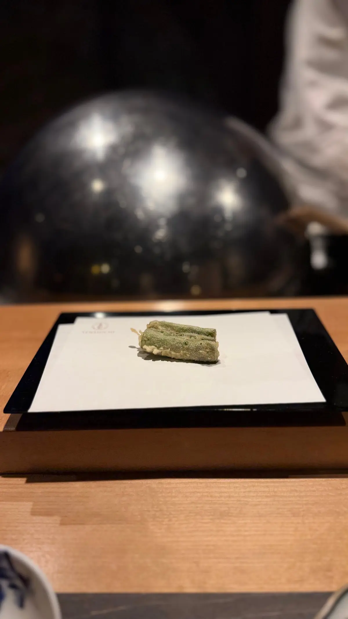 黒い台の上に置かれた、抹茶色の四角いお菓子。表面には白い粒状のものが散らばっている。背景には光沢のある黒い球体。
