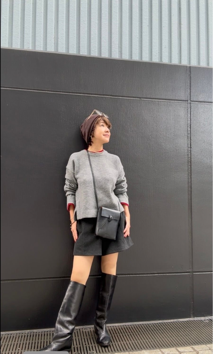 グレーのニットに黒のミニスカート、ロングブーツを合わせた女性。茶色のニット帽を被り、黒いショルダーバッグを斜め掛けしている。背景はグレーの壁。