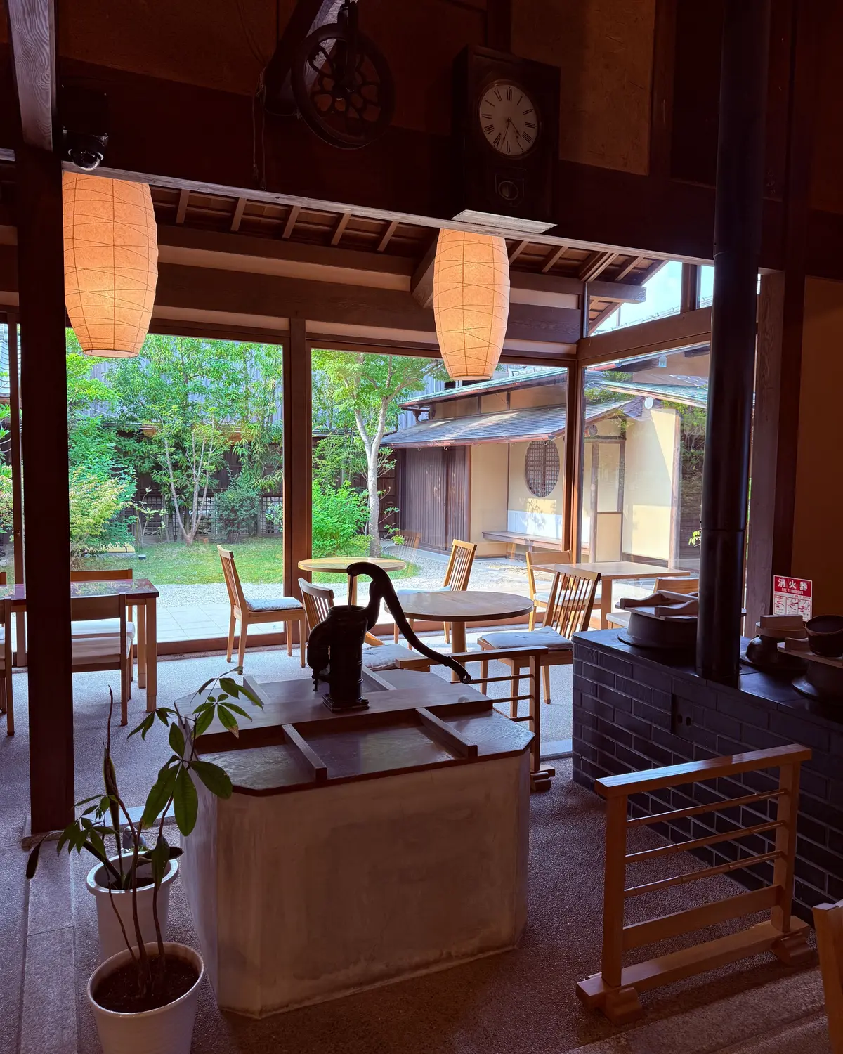 木造の茶室のような空間。障子窓の向こうには緑豊かな庭園が広がり、天井からは暖色系の灯りが吊り下げられている。中央には石の台と釜、右には鉄製の囲いのある暖炉がある。