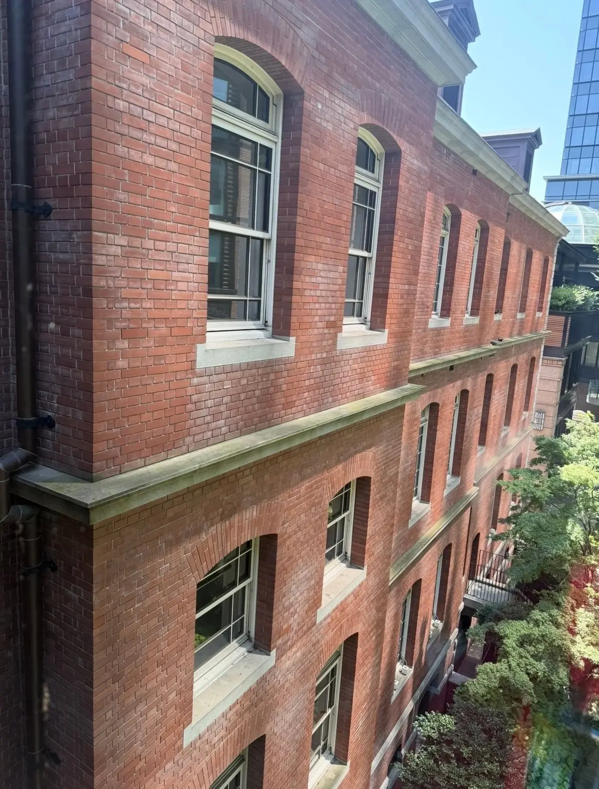 赤レンガ造りの多層ビル。アーチ型の窓と縦長の窓が規則的に並び、建物の上部には装飾的な屋根がある。右奥には高層ビルが一部見え、手前には緑の植栽が茂る。