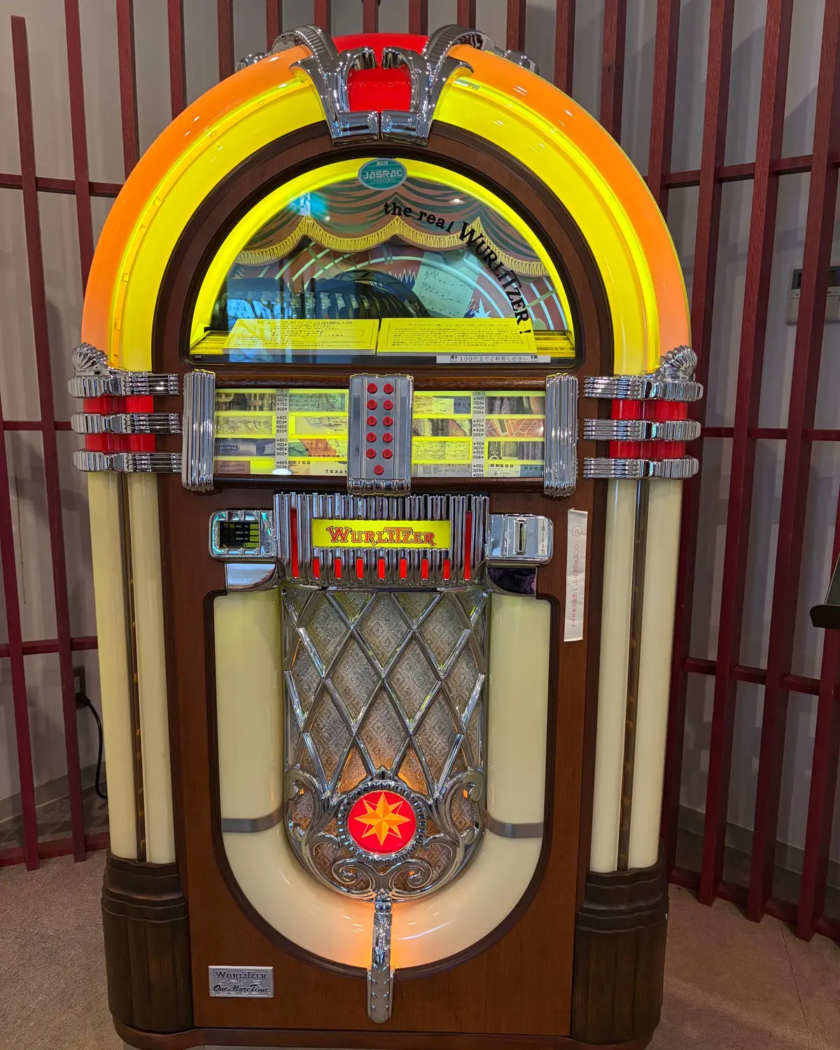 木目調のキャビネットに、黄色の光沢のある装飾が施されたWurlitzer（ワーリッツァー）のジュークボックス。中央にはレコードジャケットのイラストが描かれ、ボタン式の選曲パネルが配置されている。