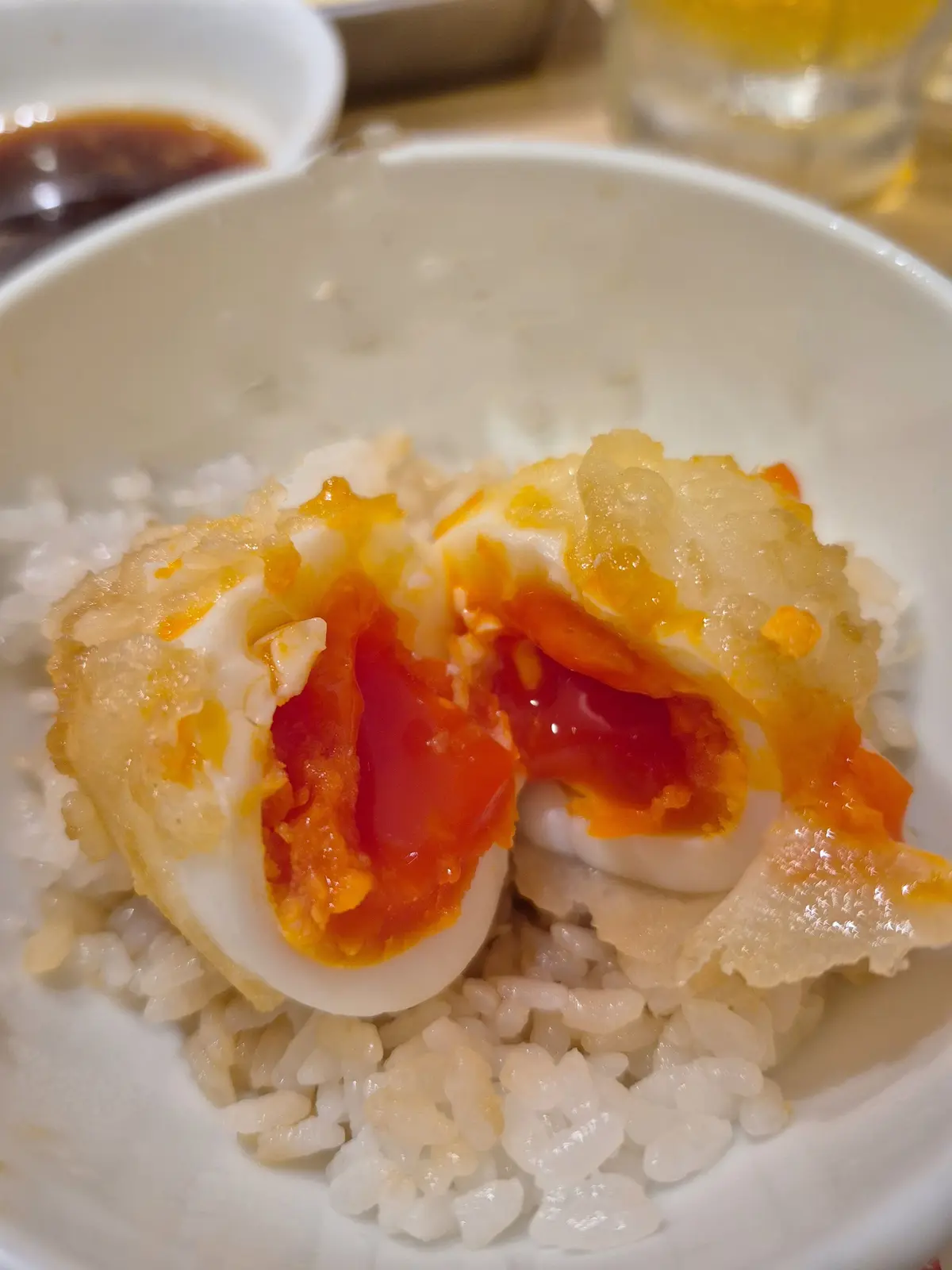 白米の上に、揚げたての卵が乗った丼。卵は半分割れており、中からオレンジ色の卵黄が染み出している。丼の脇には、醤油のような液体が入った小皿が置かれている。